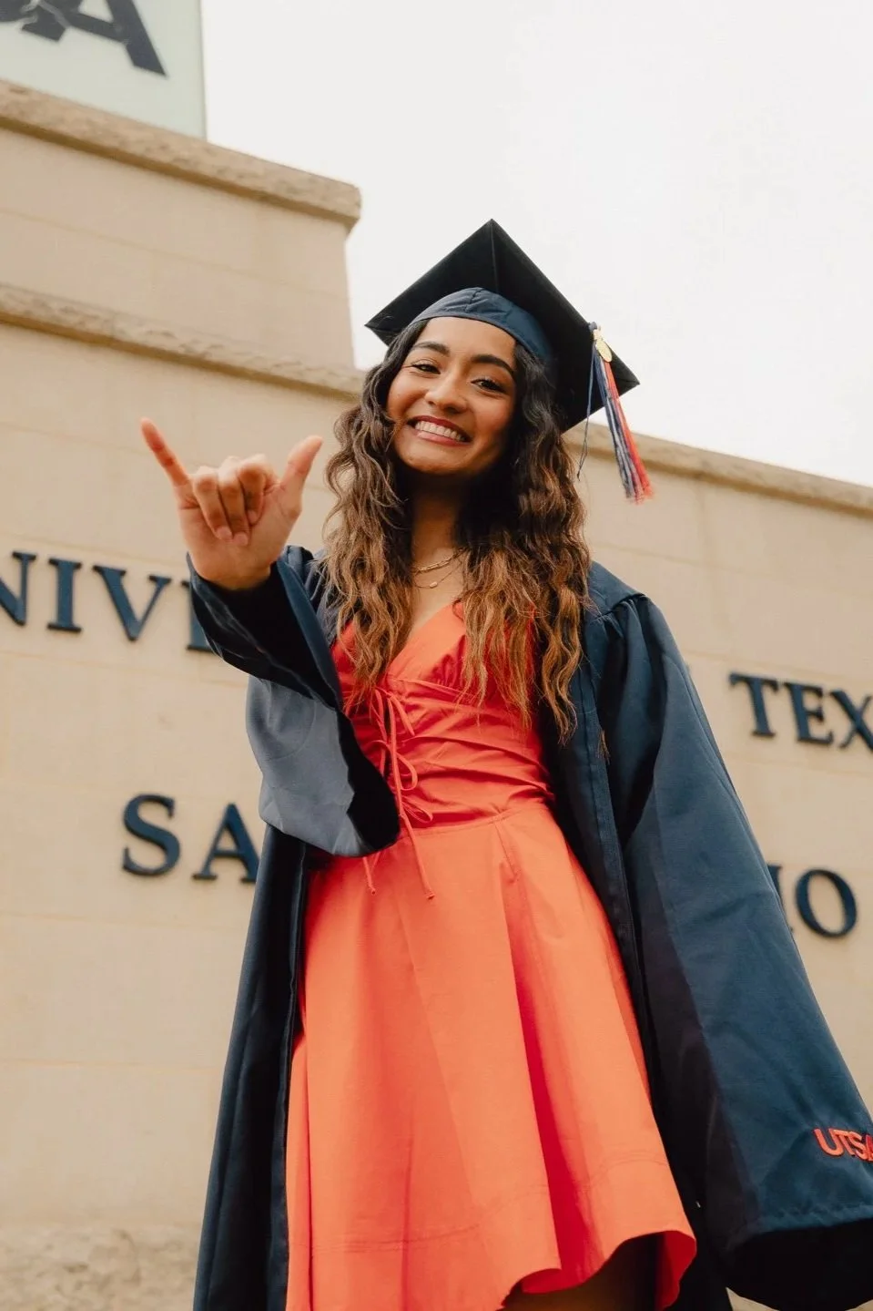 A young woman in a graduation cap and gown smiling and showing a shaka sign in front of a university sign.