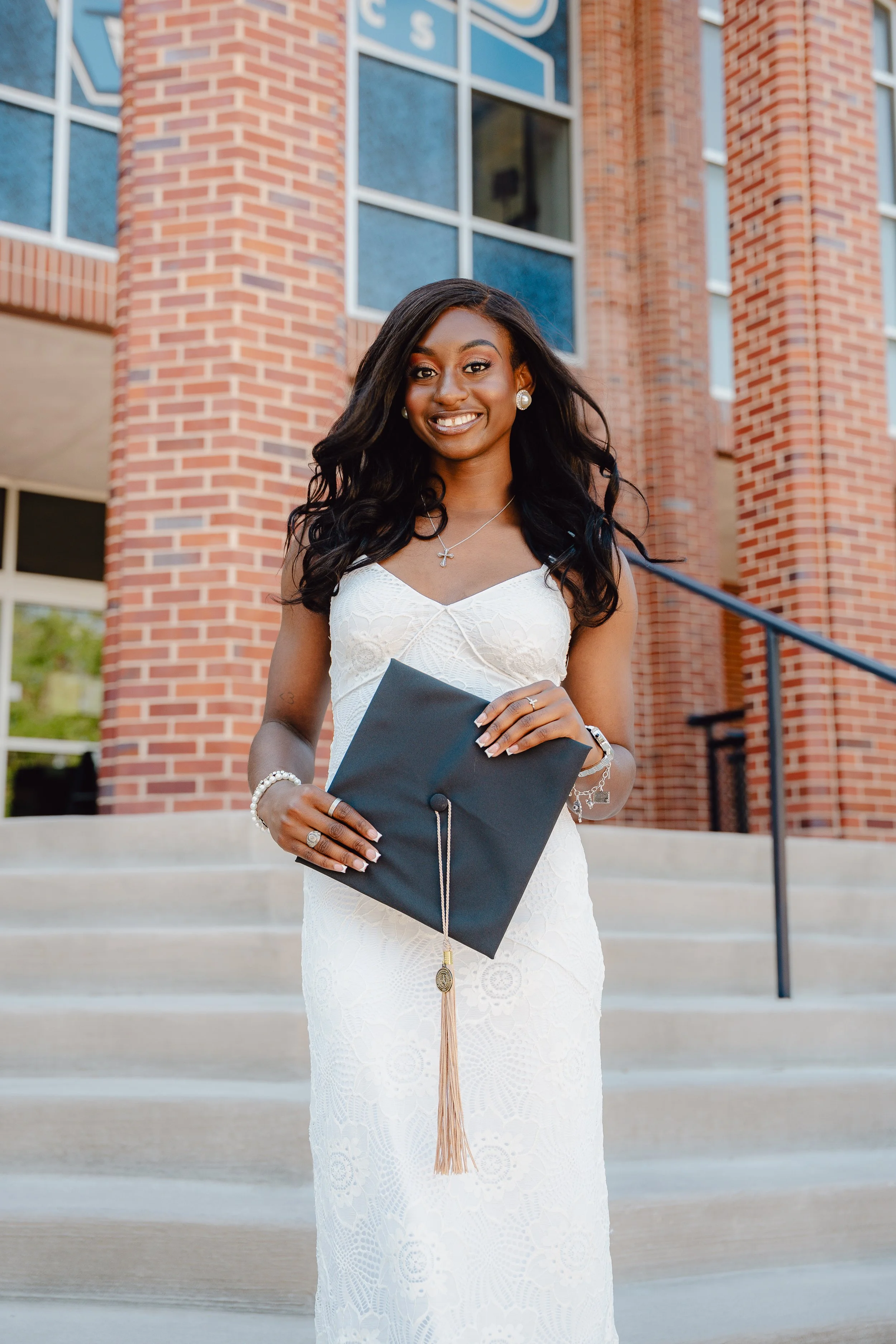 A young woman dressed in a white dress holding a diploma in front of a brick building with large windows, celebrating graduation.