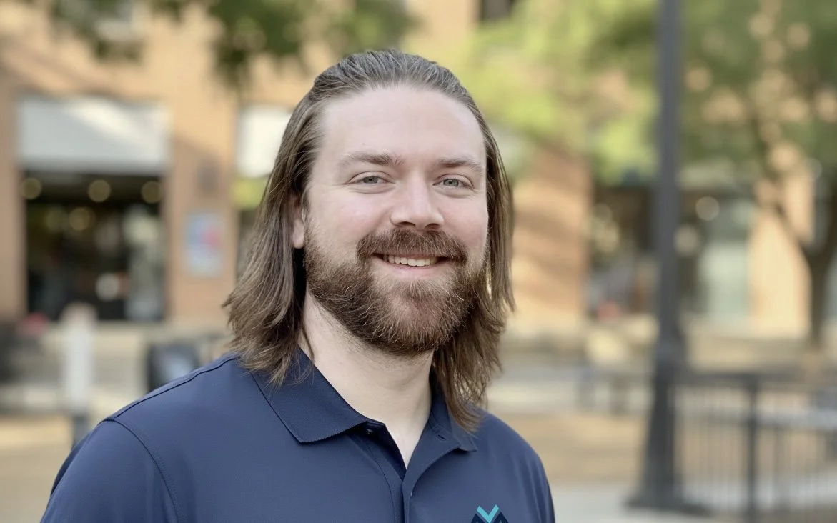 Portrait of a young man with shoulder-length brown hair, beard, wearing a navy polo shirt, standing outdoors in a park with trees and buildings in the background.