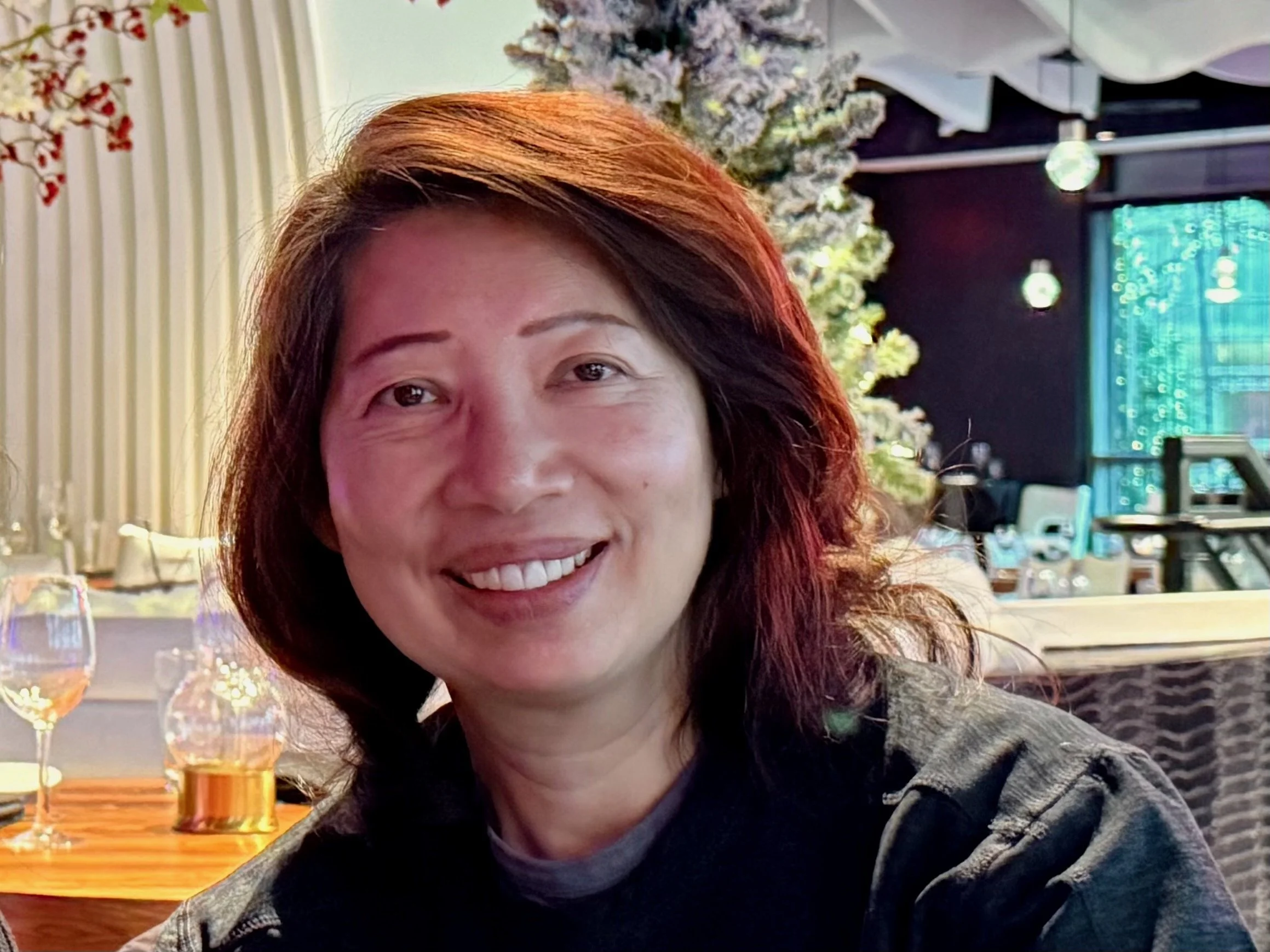 A woman with curly dark hair and hoop earrings, wearing a beige sleeveless top and brown high-waisted pants, sitting on a cushioned bench in a modern interior with wooden wall paneling and soft lighting.