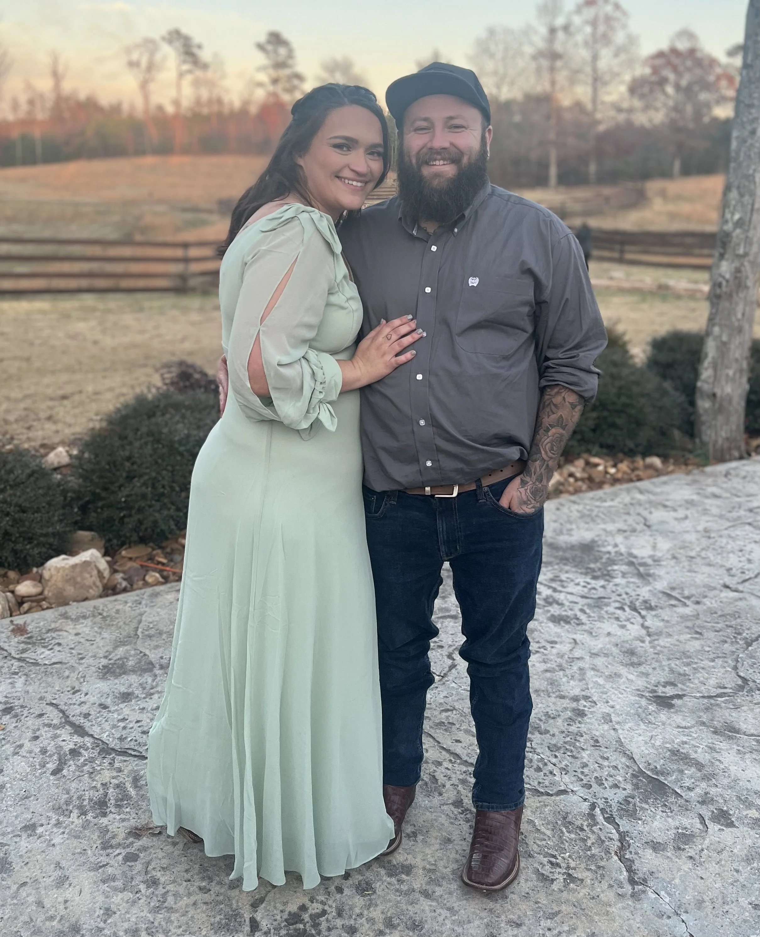 A woman and a man standing close together outdoors on a stone-paved area with a wooden fence and trees in the background during sunset. The woman is wearing a light green dress with cutout sleeves, and the man is dressed in a dark gray button-up shirt, jeans, and brown boots, with a black cap and a long beard, both smiling at the camera.