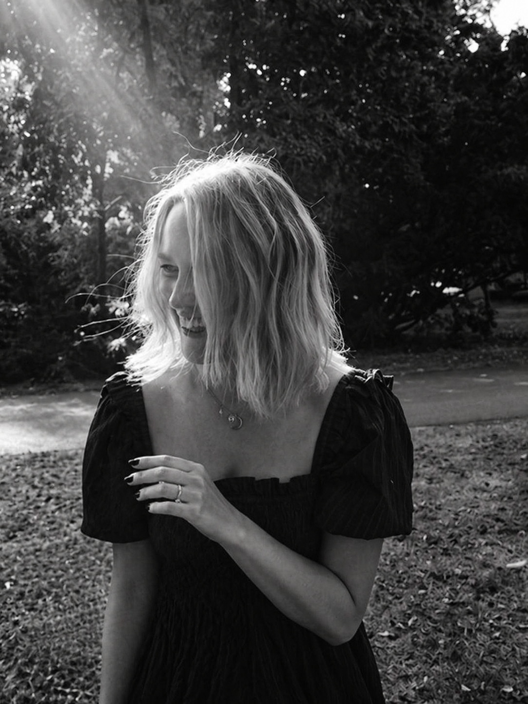 A smiling woman with shoulder-length wavy blonde hair standing outdoors in sunlight, wearing a dark dress with puffed sleeves, a necklace, and rings, with trees and a grassy area in the background.