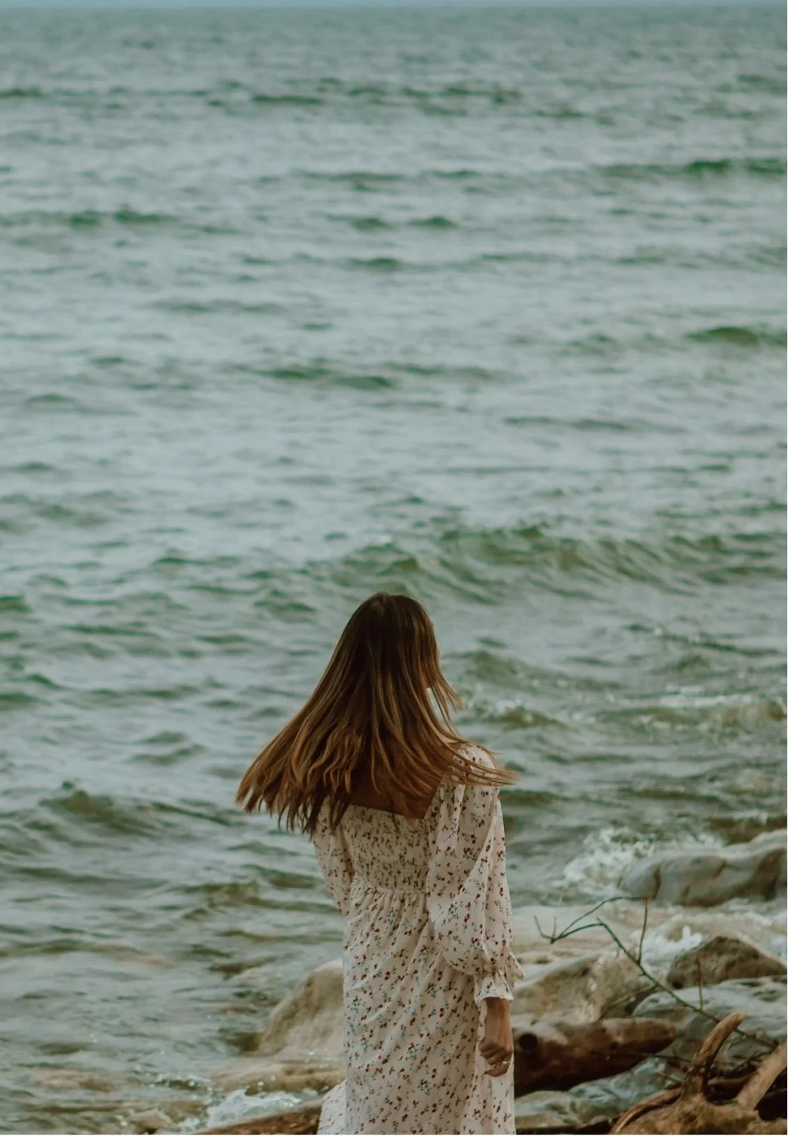 A woman with long brown hair wearing a white floral dress standing on rocks at the edge of the ocean with waves, facing away from the camera.