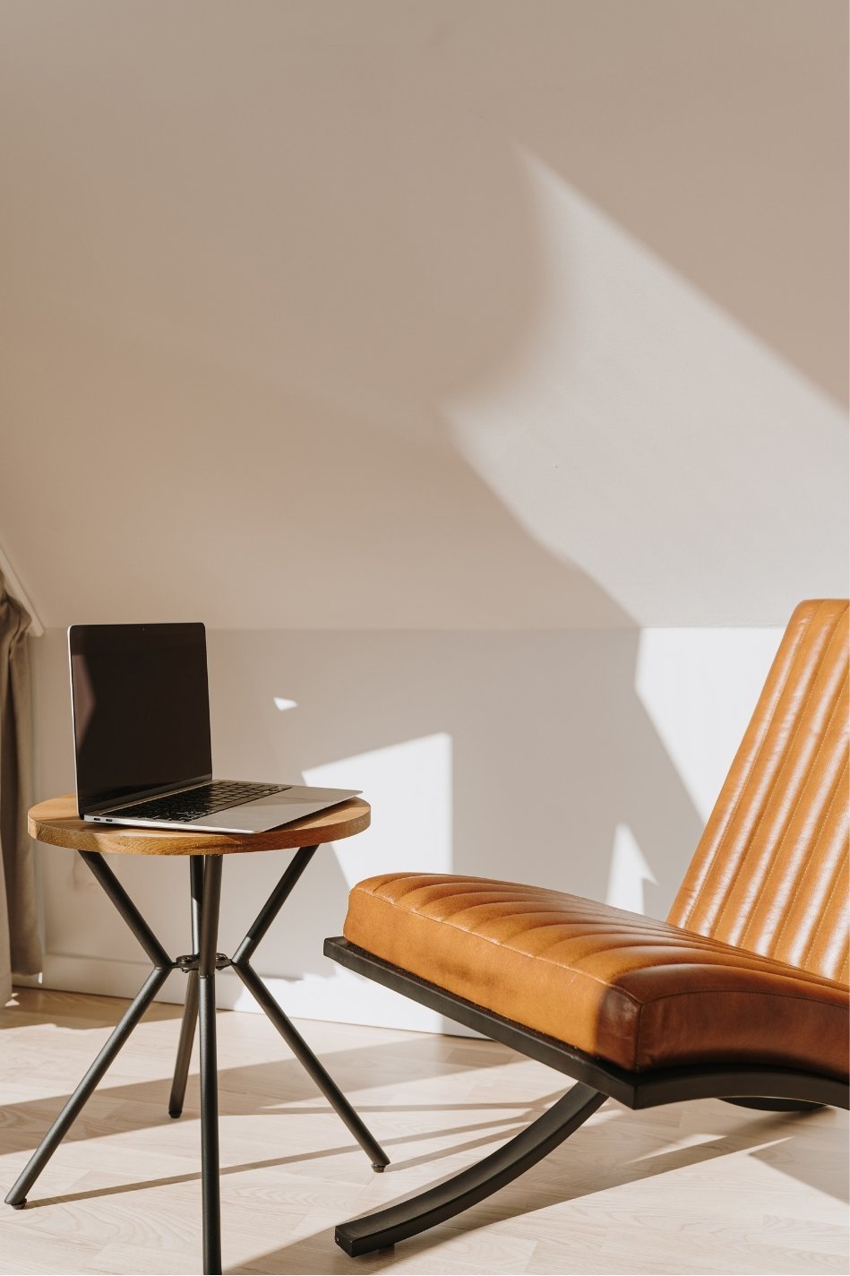 Modern room with a small round wooden table holding a black open laptop, a mid-century style orange leather lounge chair with black base, on light wood flooring, with sunlight casting geometric shadows on the wall.