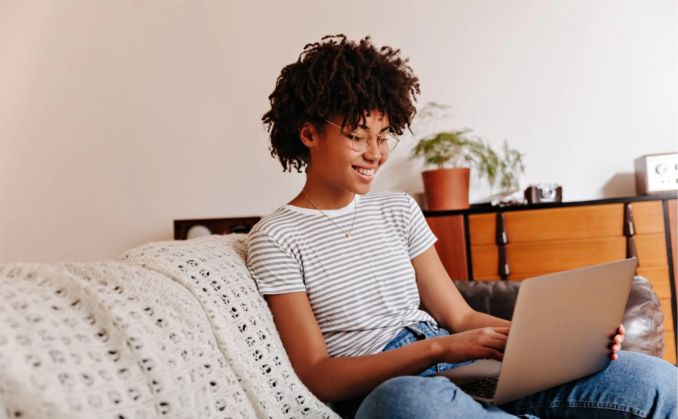 A young woman with glasses and curly hair, wearing a striped shirt, sitting on a cozy sofa with a knitted blanket, looking at her laptop and smiling.