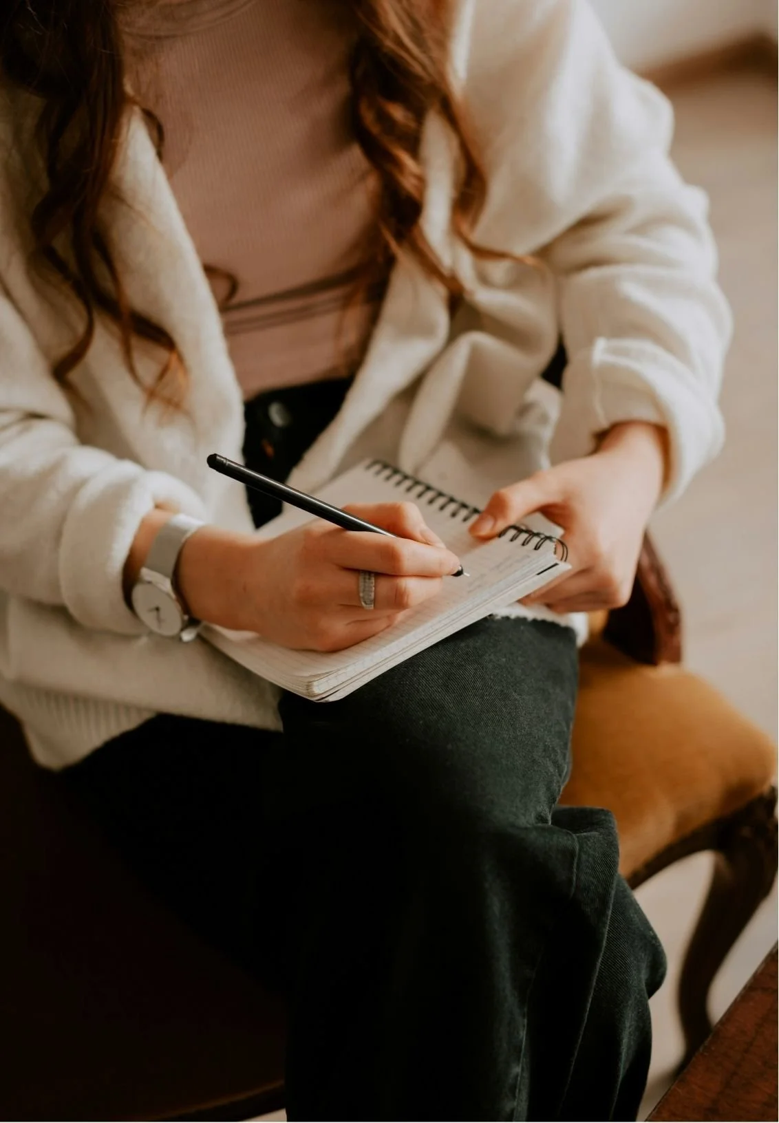 A woman sitting on a chair writing in a small spiral notebook with a black pen, wearing a beige sweater, black pants, and a white watch on her left wrist.