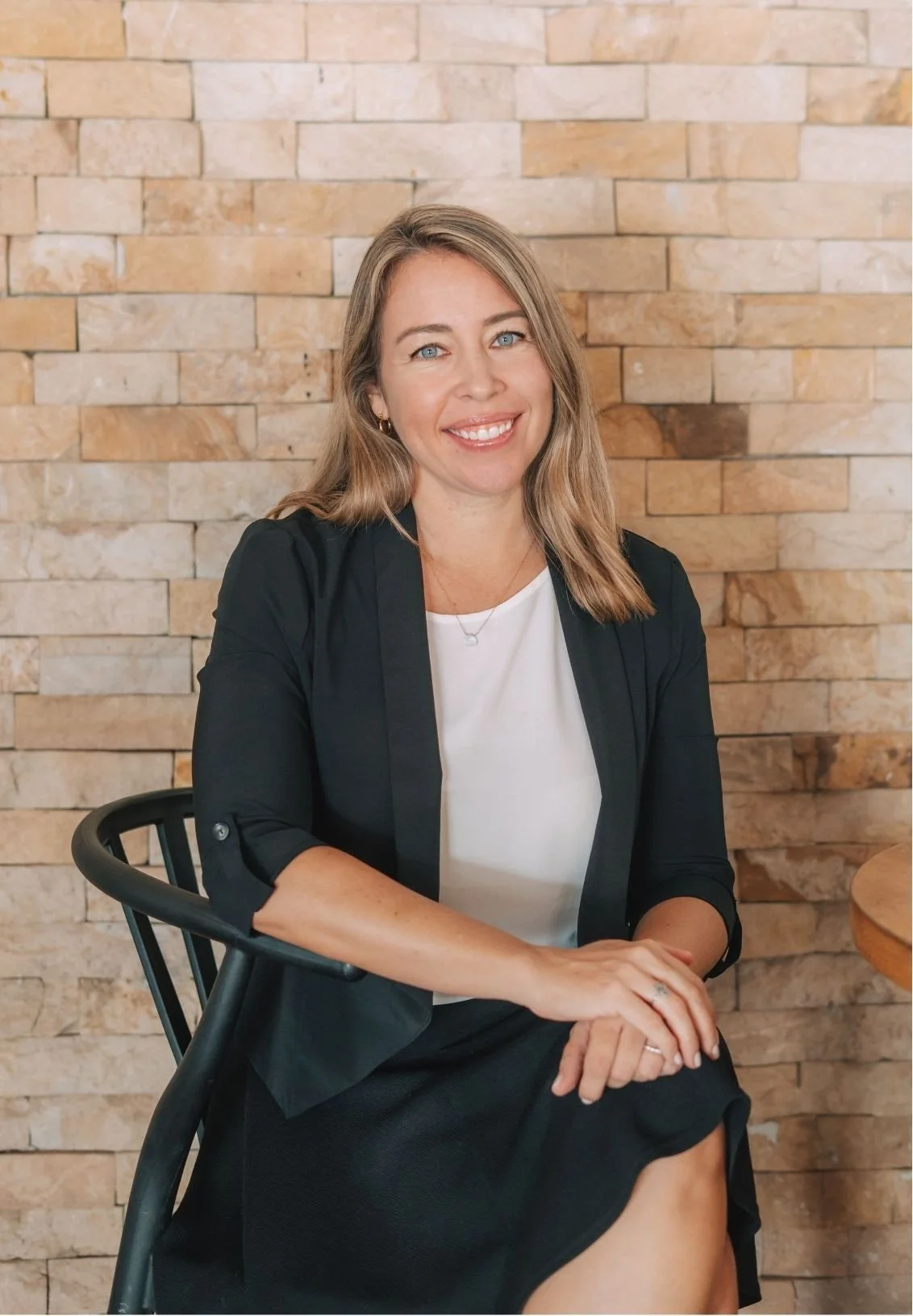 Dr. Ashley Lynch, EMDR therapist in CA, with shoulder-length brown hair, blue eyes, wearing a black blazer over a white top, sitting on a black chair in front of a beige brick wall, smiling at the camera.