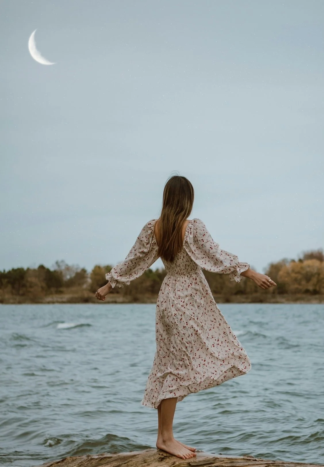 A woman in a floral dress standing on a rock by a lake at dusk, facing away from the camera, with a crescent moon and starry sky overhead.