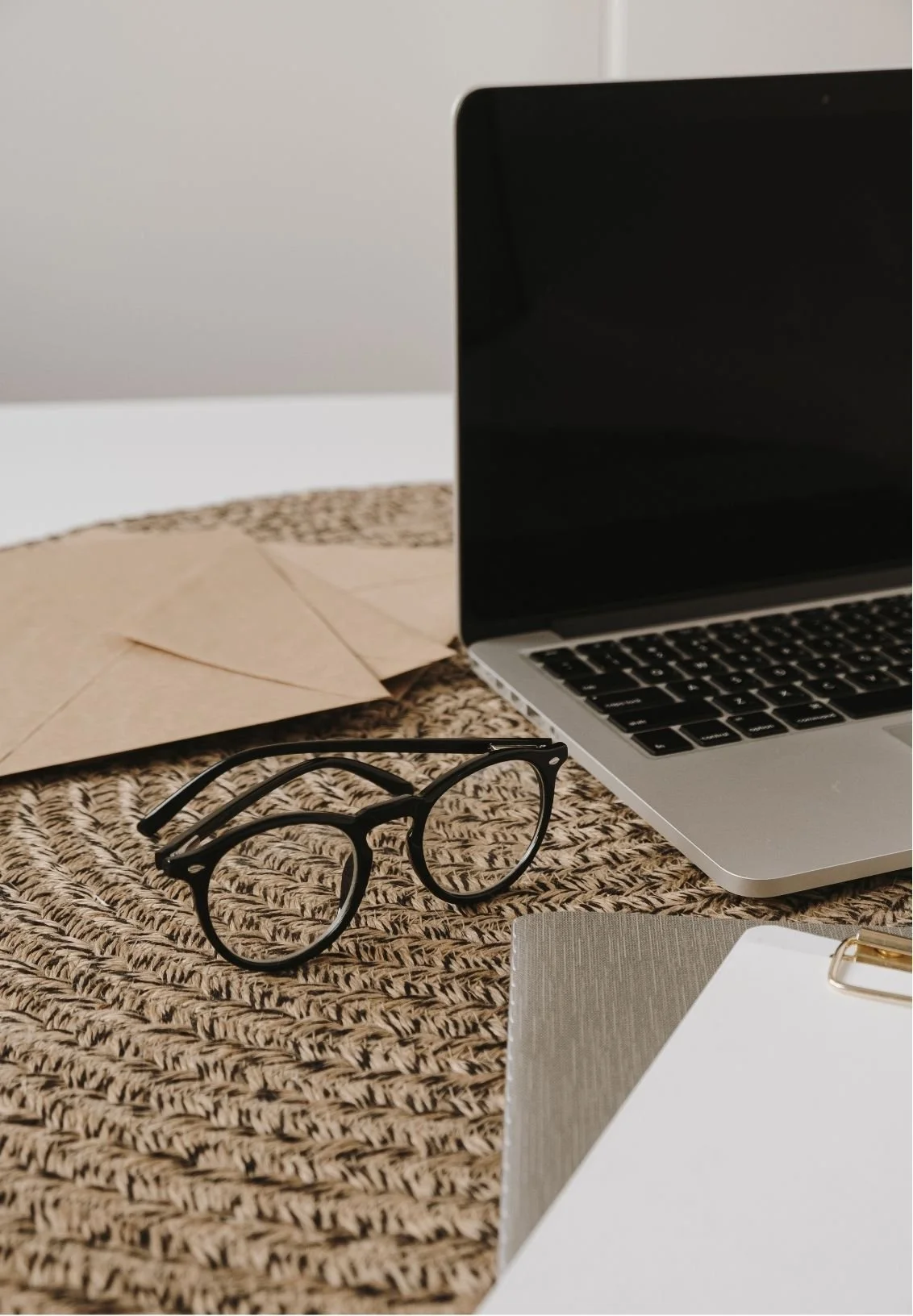 A pair of black eyeglasses, a laptop, brown envelopes, and a notepad with a paper clip on a woven table mat.