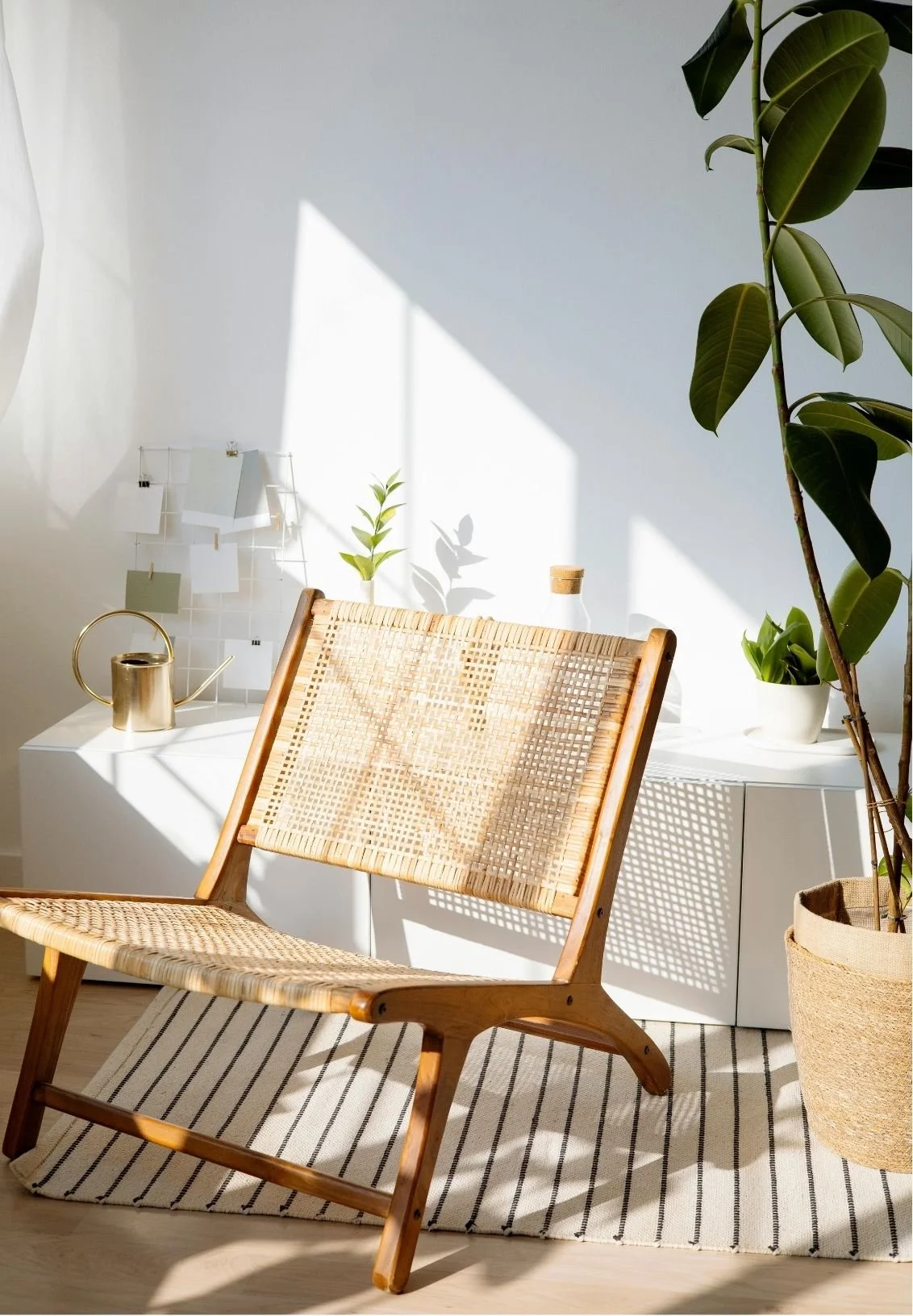A cozy corner with a wooden and woven chair, a white side table with potted plants, a gold watering can, and a small grid with paper in natural sunlight.