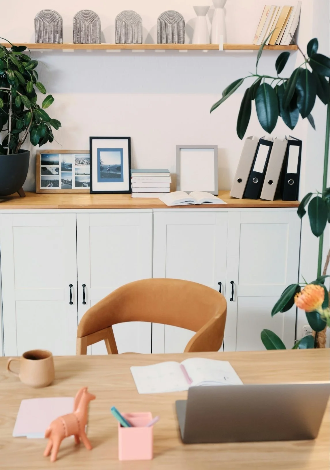A home office or study space with a wooden table, a leather chair, a white storage cabinet, and a shelf with decorative items, books, and photo frames. There are indoor plants on both sides of the image.