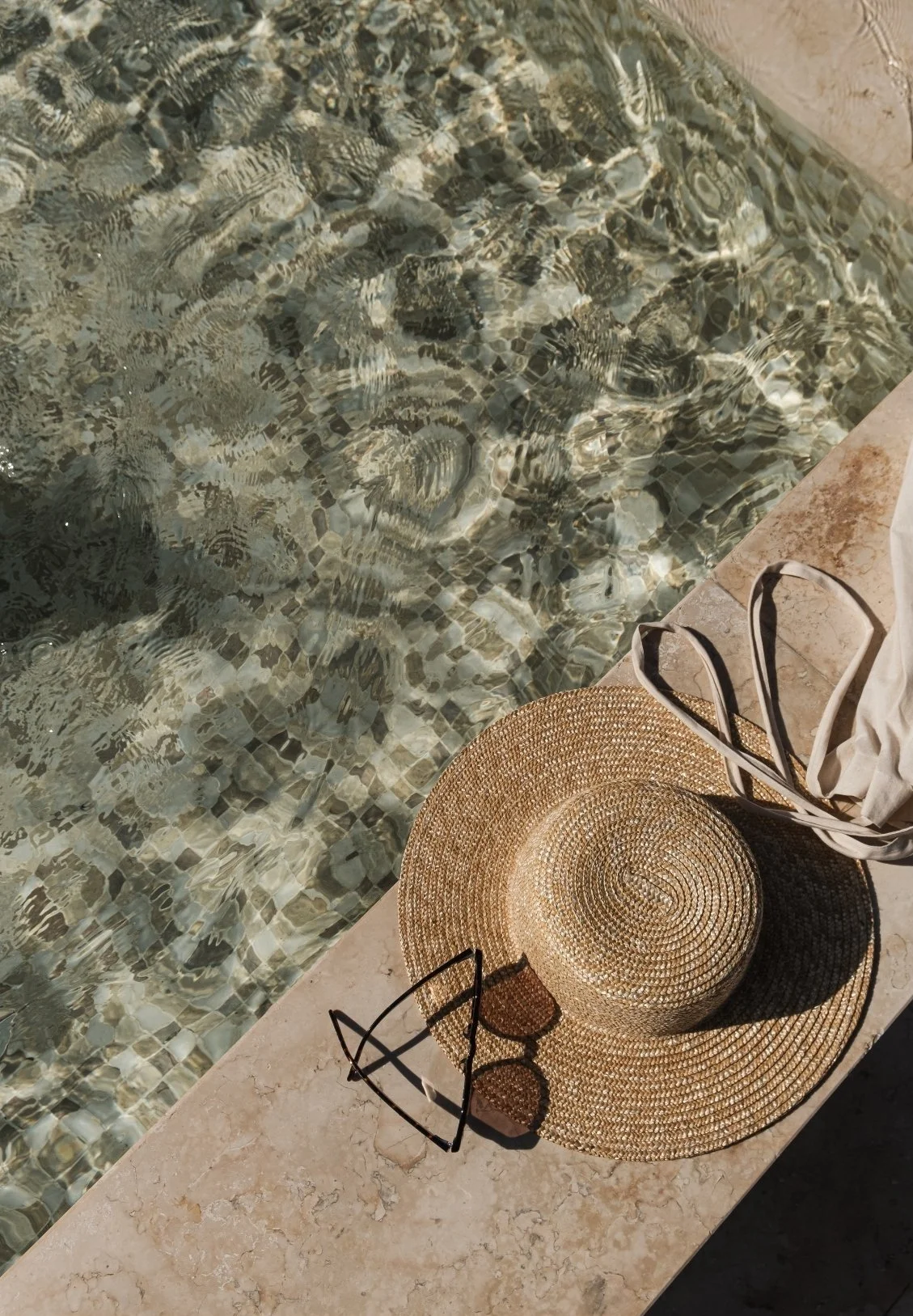A close-up view of a swimming pool with clear water, a straw hat, sunglasses, a cloth bag, and a pair of sandals on a tiled poolside surface.