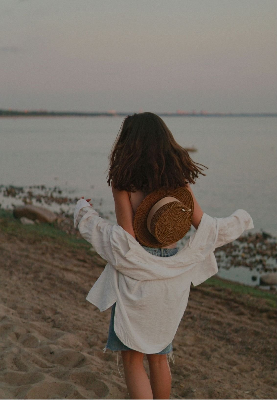 A girl with brown hair standing on a beach by the water at sunset, wearing a white shirt, denim shorts, and holding a straw hat on her back.