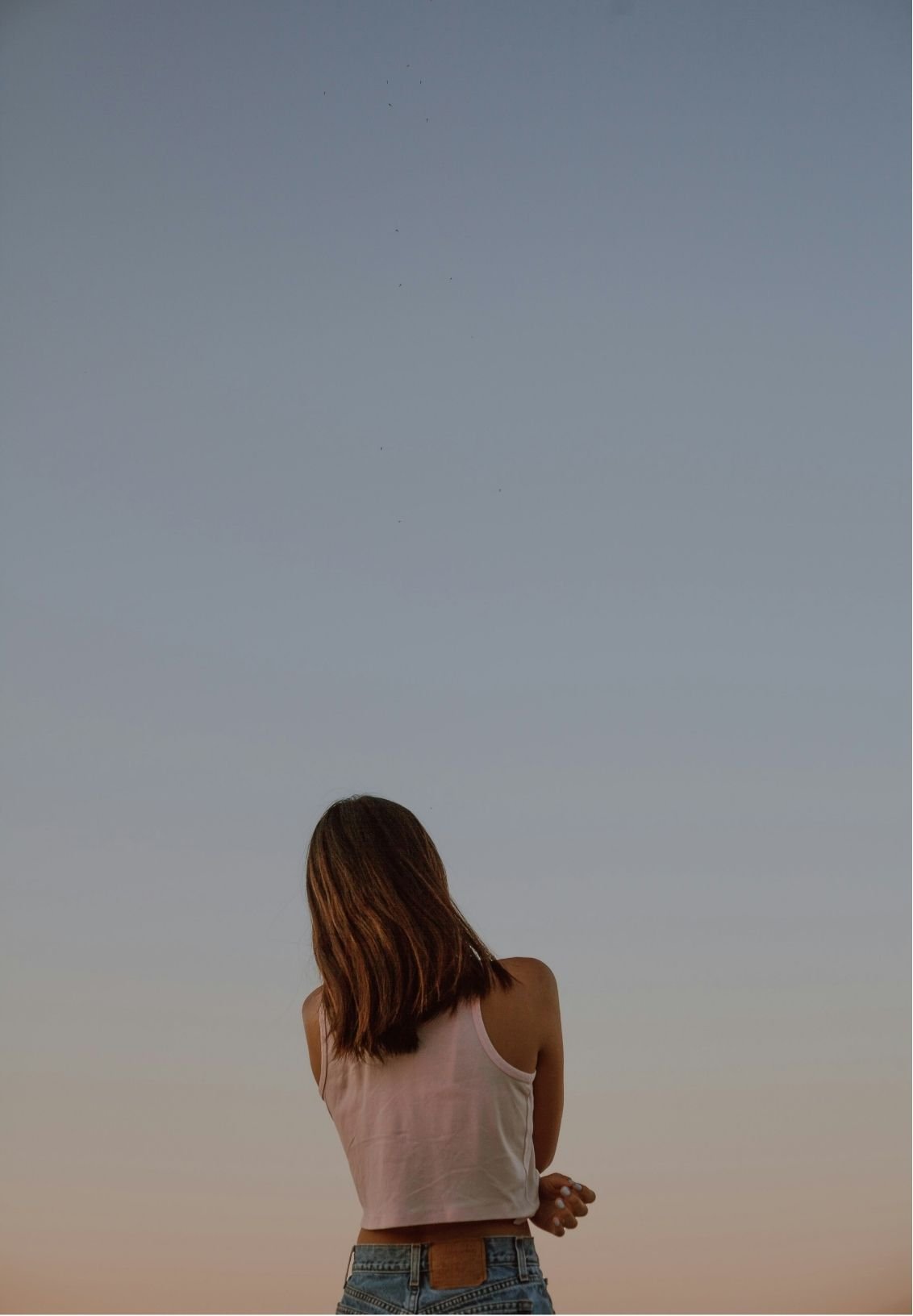 A woman with brown hair wearing a white sleeveless top and jeans stands outdoors at dusk, facing away, with the sky behind her.