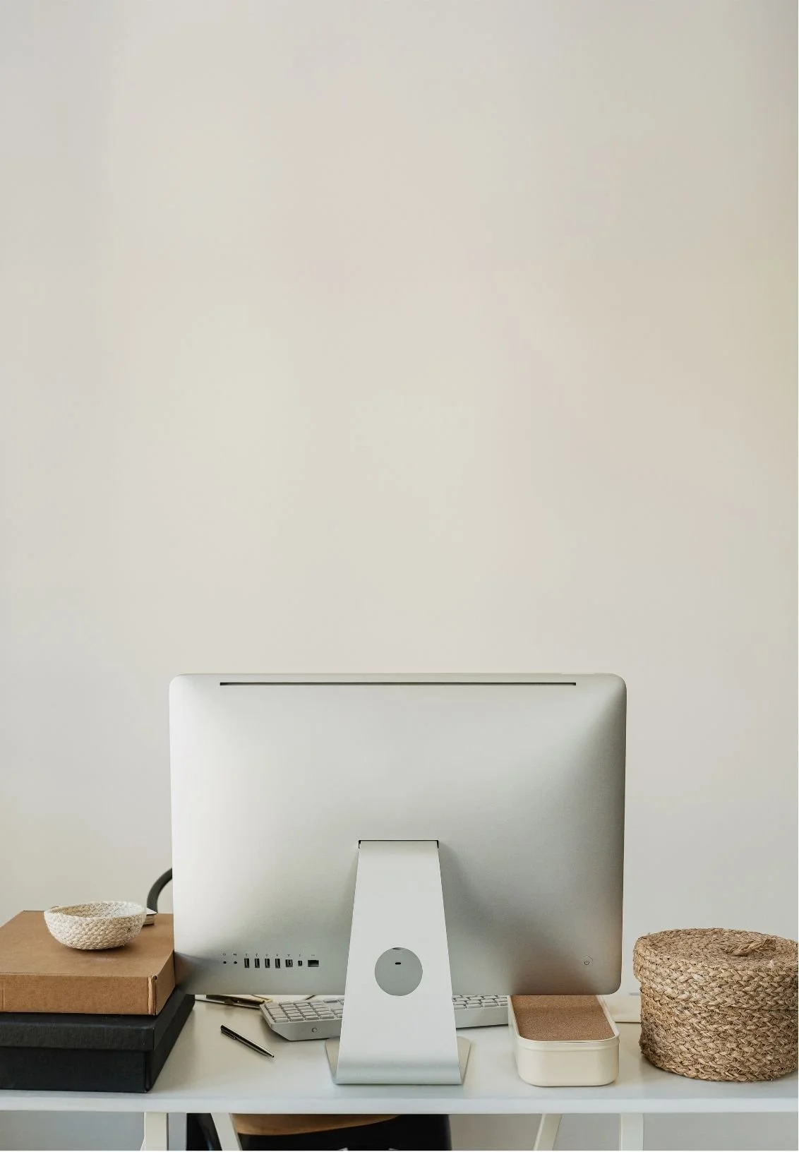 A minimalist white desk setup with a computer monitor, wireless keyboard, and mouse. To the left, there are folders and a small woven bowl. On the right, there is a woven basket and a container with a brown lid. The background is a plain, light-colored wall.