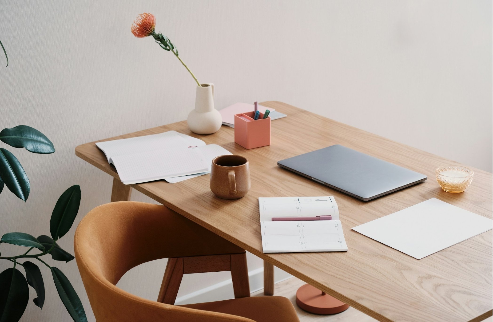 A wooden desk with a closed laptop, an open planner, a pink pen holder with pens, a white notebook, a brown mug, and a small glass bowl. There is a white vase with a single orange flower, and a large green plant with broad leaves nearby. A beige chair is in front of the desk, and part of a white wall is visible in the background.