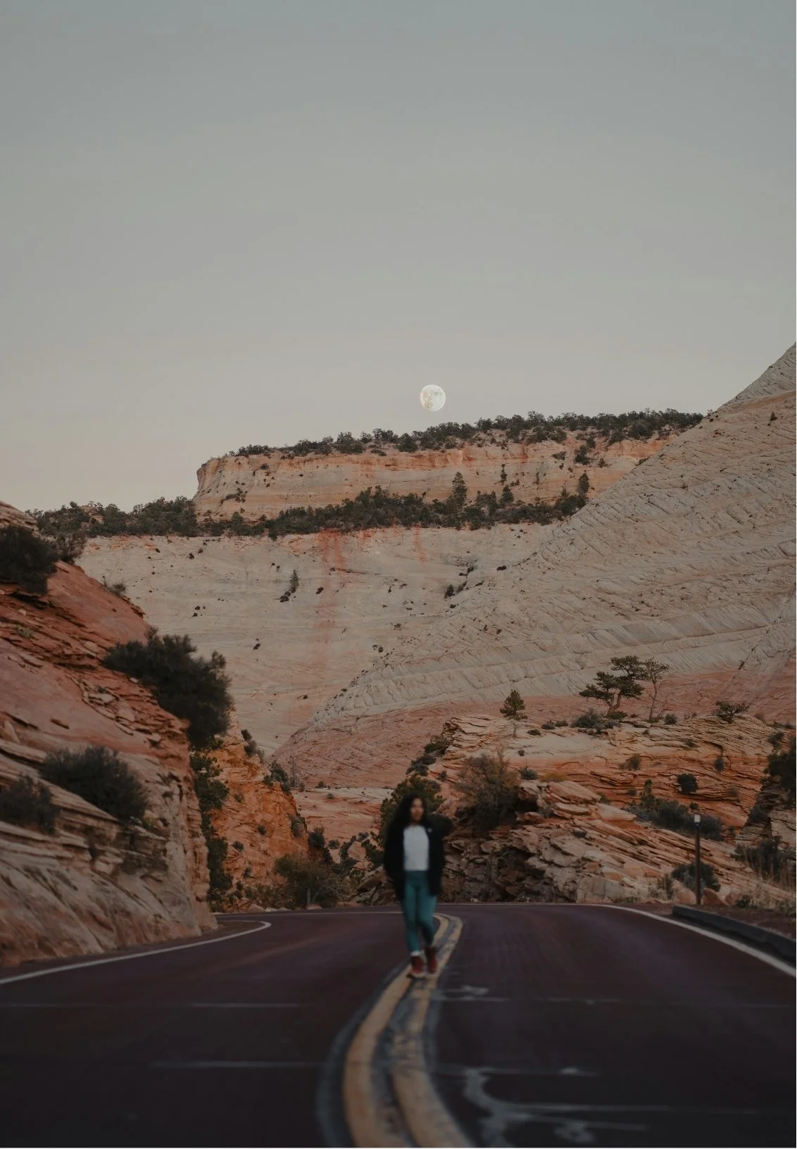 A person walking on a winding road through a desert canyon landscape with rocky cliffs and sparse vegetation, under a clear sky with the moon visible.