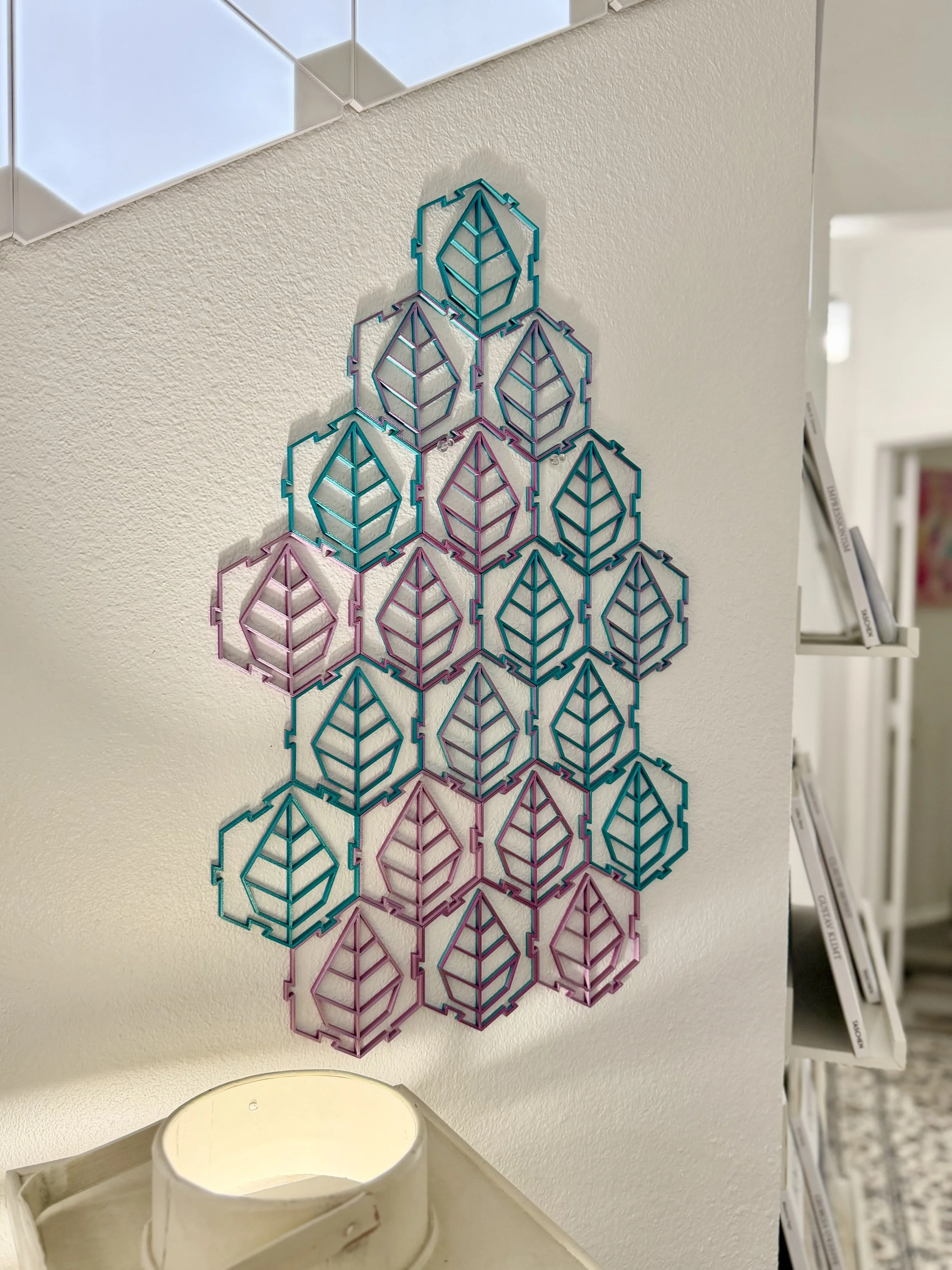 Colorful geometric wall art piece made of interconnected hexagon and leaf-shaped wireframes hanging on a beige textured wall in a room with shelves holding books.