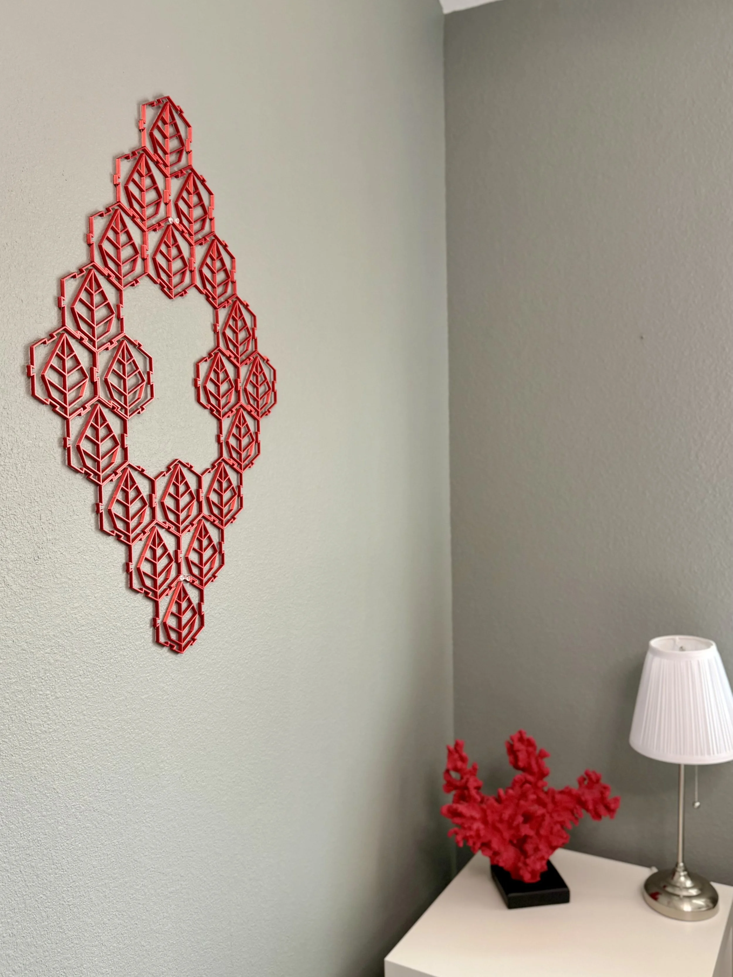 Decorative red wall hanging in a geometric leaf pattern on a light green wall, with a white side table below holding a black vase with red flowers, and a white table lamp.