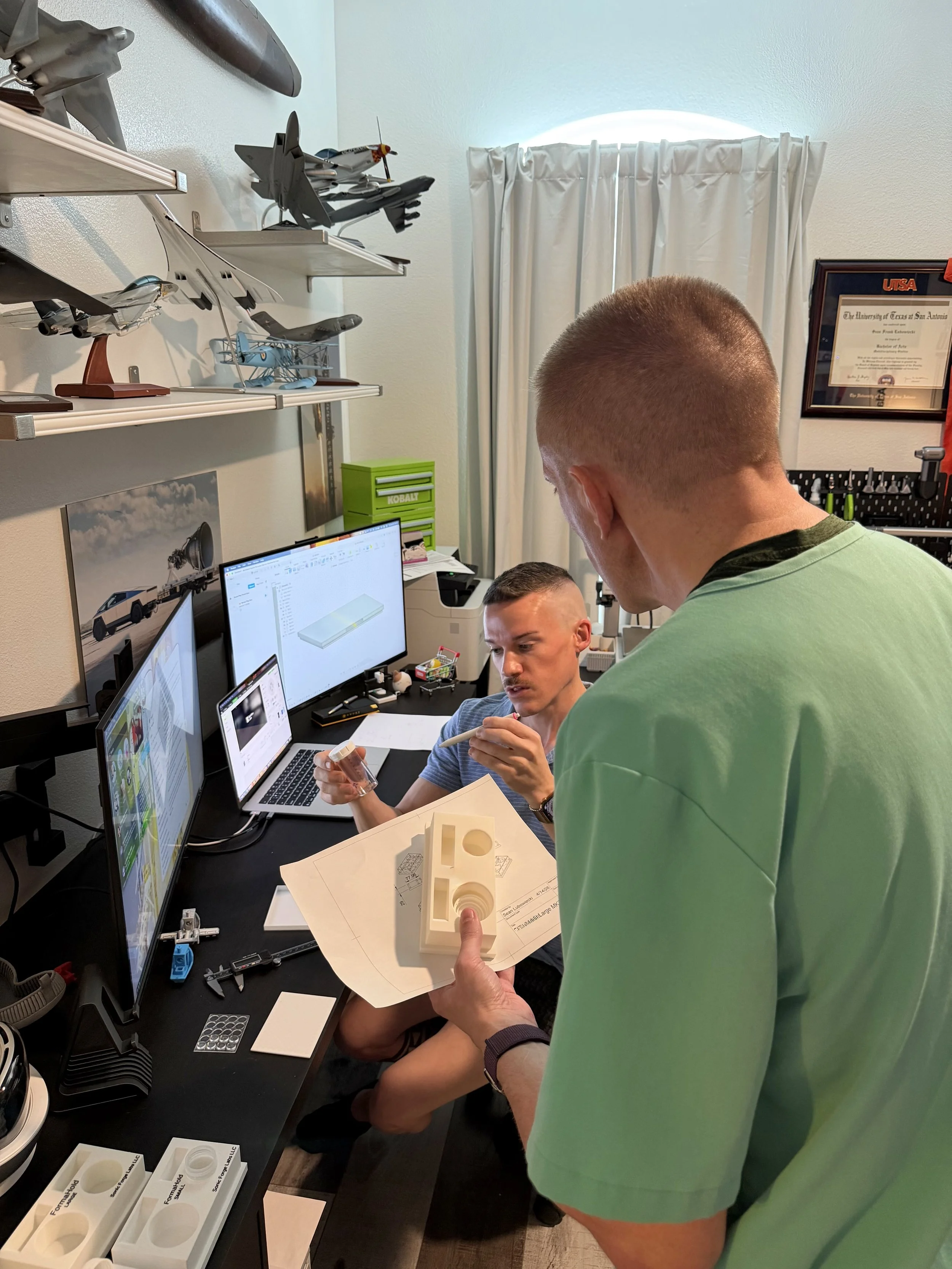 Two men working in a room with multiple model airplanes on shelves and a framed diploma on the wall. One man is holding a model part and a blueprint, while the other is looking at the computer screens.