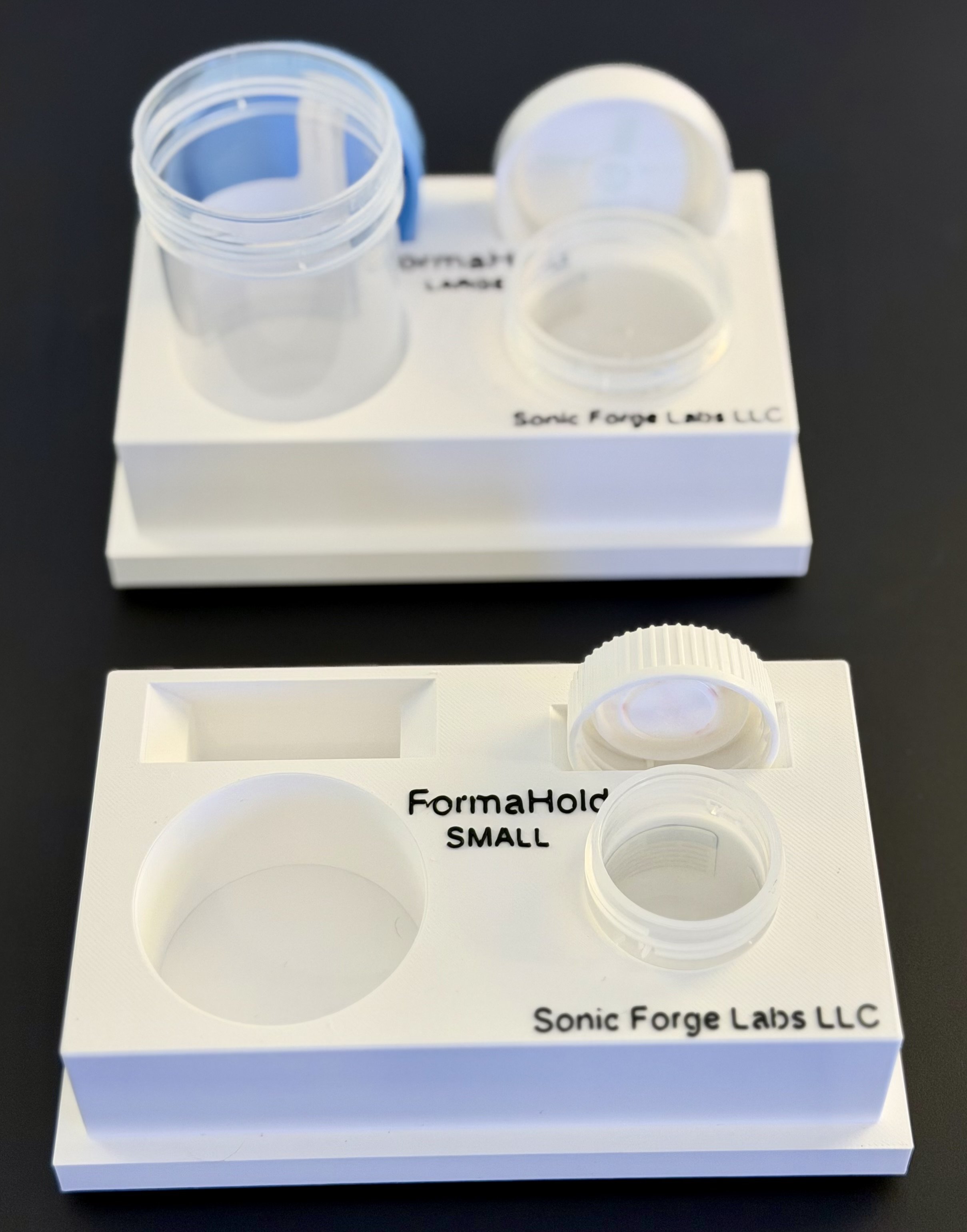 Two white plastic holders with empty circular slots, labeled 'FormaHold SMALL' and 'Sonic Forge Labs LLC,' placed on a black surface, with one holder partially stacked on top of the other.