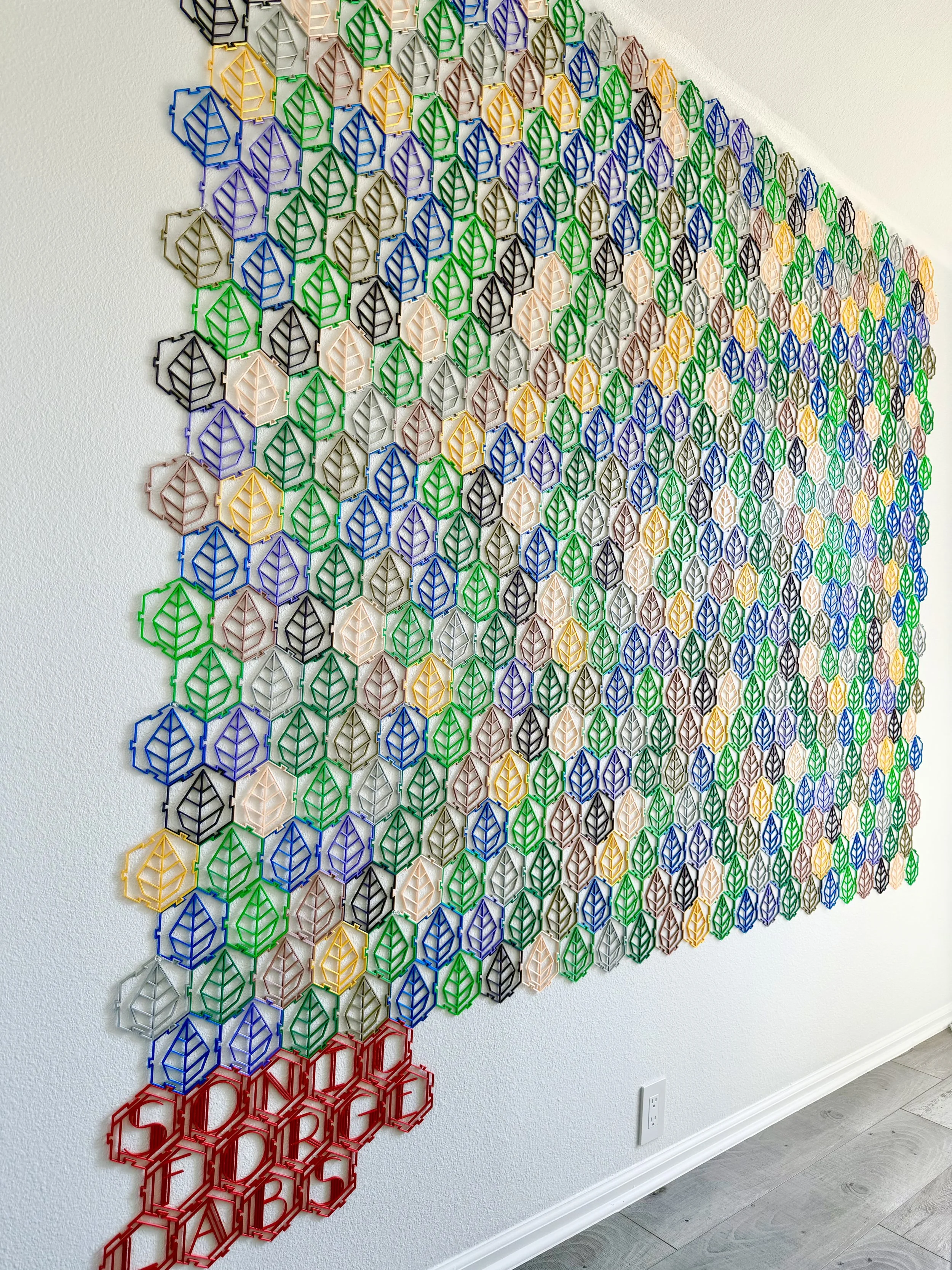 A wall display of colorful geometric artwork composed of interconnected plastic hexagons, arranged in a cascading pattern from black, green, purple, silver, yellow, blue, beige, until red colors, covering a large section of the wall.