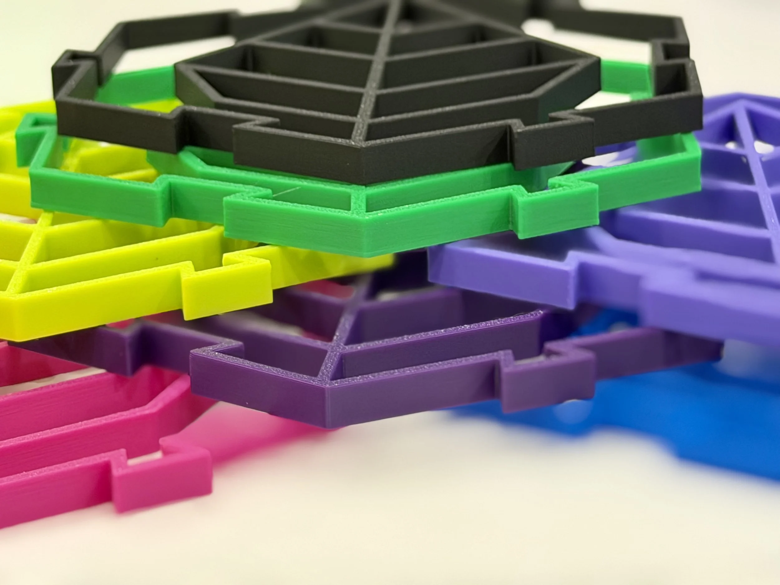 Close-up of colorful plastic geometric shapes stacked in layers, including pink, purple, yellow, green, and black, on a white surface.