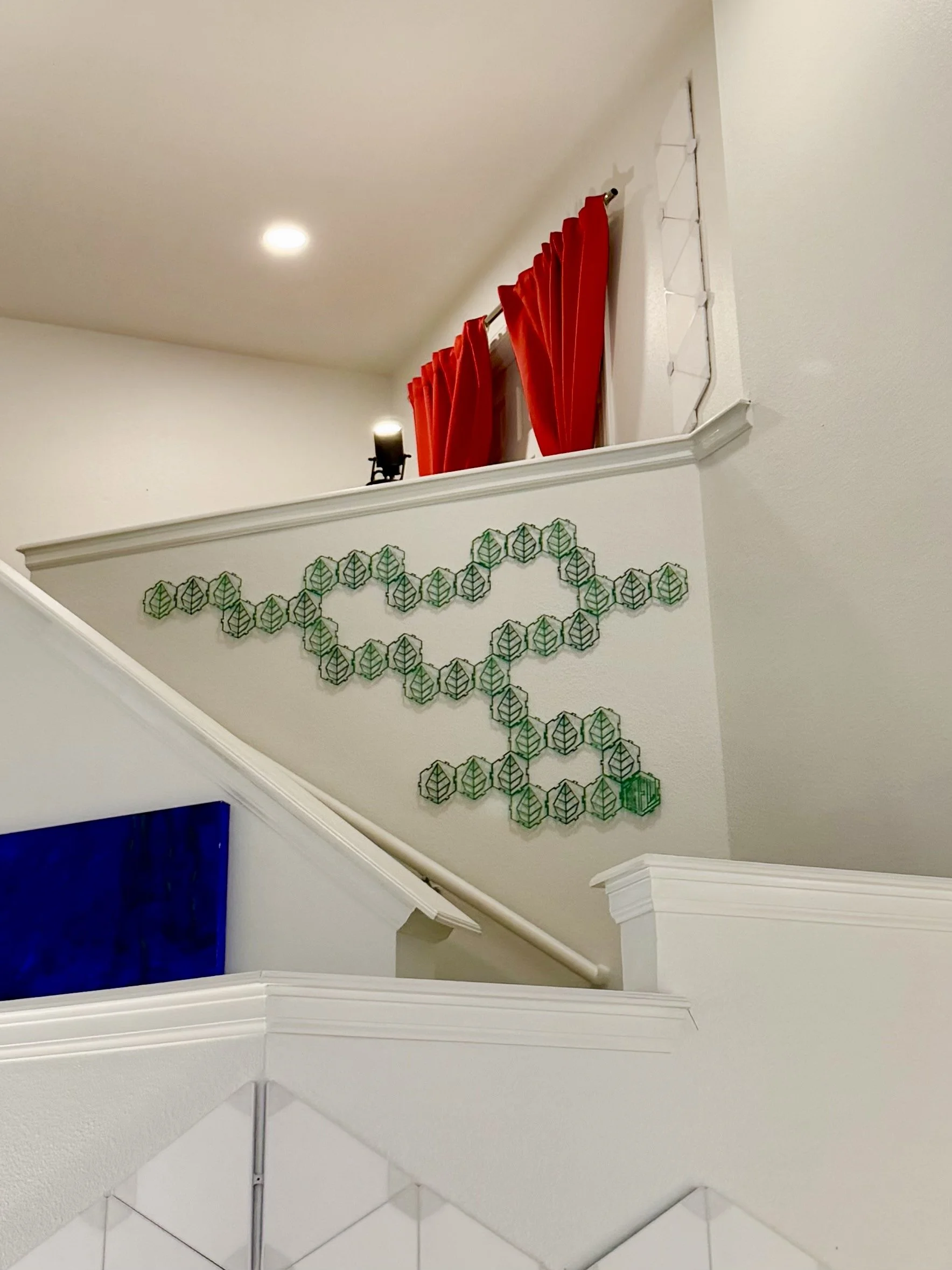 Interior view of a staircase with white rails, wall decorations resembling chlorophyll molecules, red curtains on a higher window, and a small black lamp on the upper wall.
