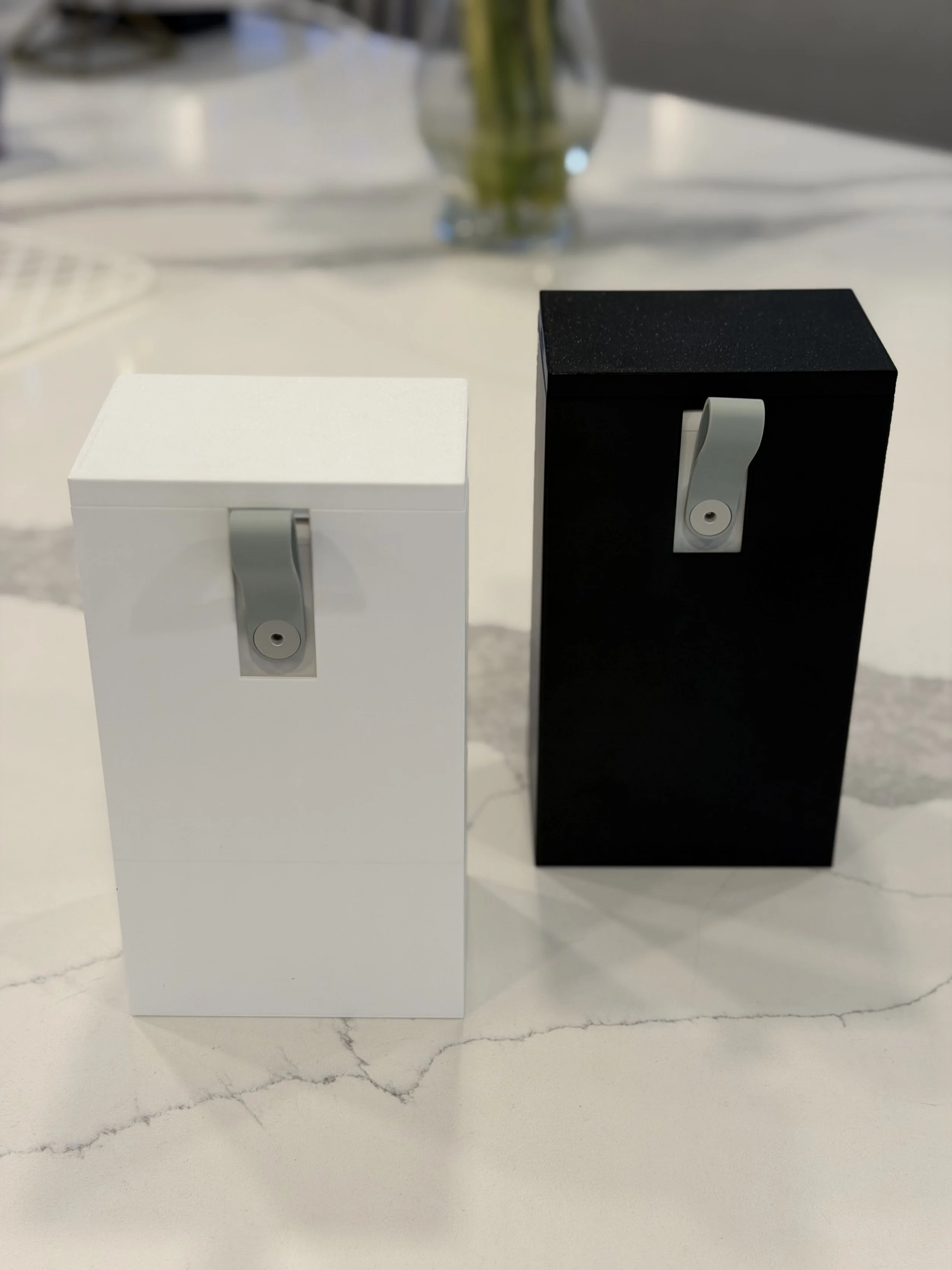 Two rectangular boxes, one white and one black, with dispensers attached to their fronts, placed on a white marble surface.