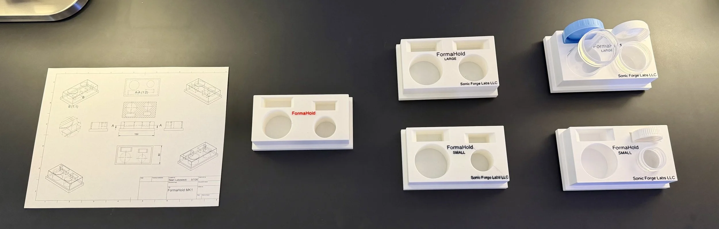 Various sizes of white FormaHold molds labeled Large, Small, and with evolving designs, placed on a black table with a technical drawing on the left.