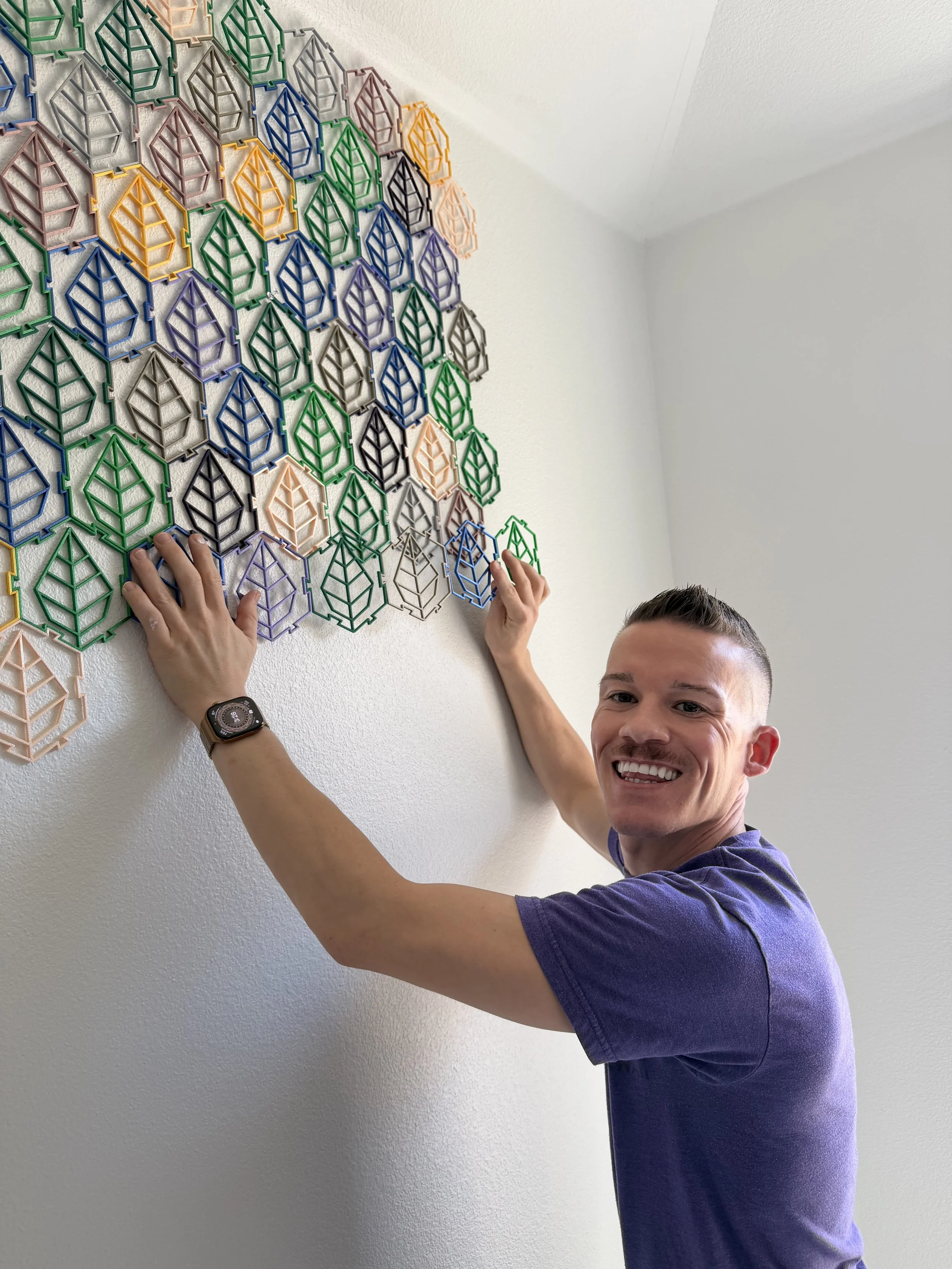 A smiling man with short hair, arranging or adjusting colorful plastic or metal geometric wall art pieces with his hands.