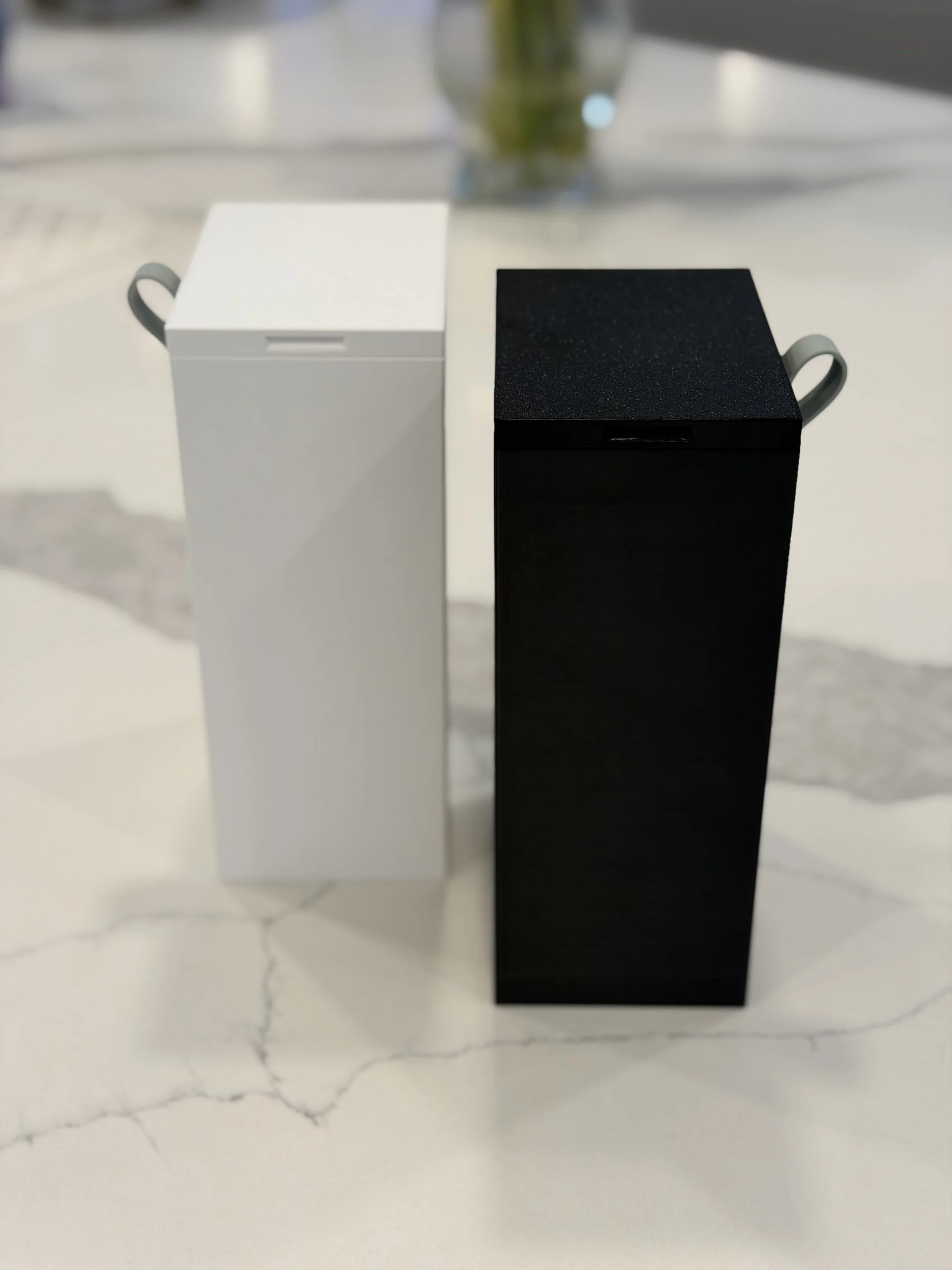 Two rectangular boxes, one white and one black, are placed vertically on a marble surface, with the white box on the left and the black box on the right.