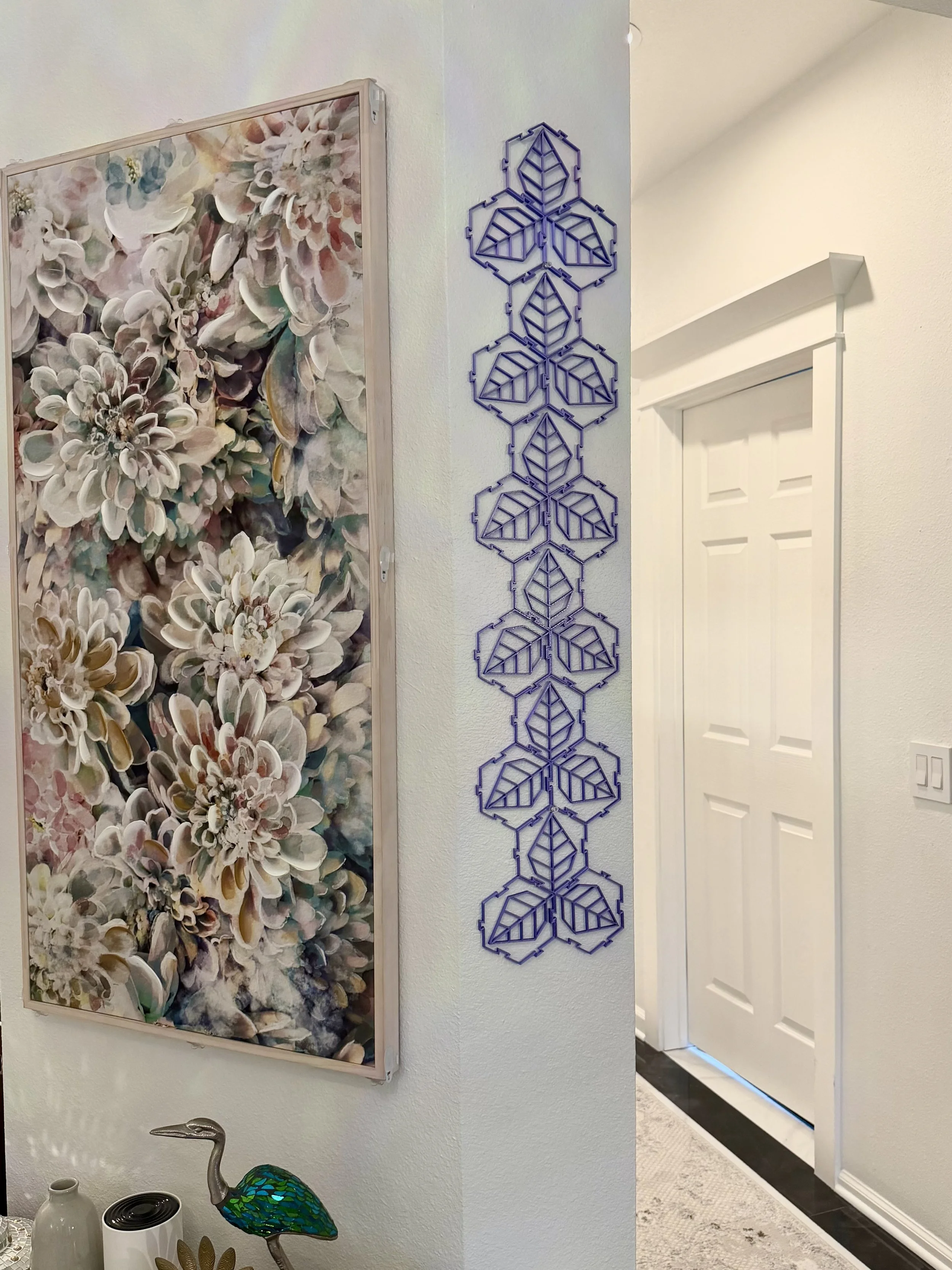 Interior of a home with floral artwork on the wall, decorative blue metal wall art with geometric leaves, a white door, and a small table with a mosaic peacock sculpture and some vases.