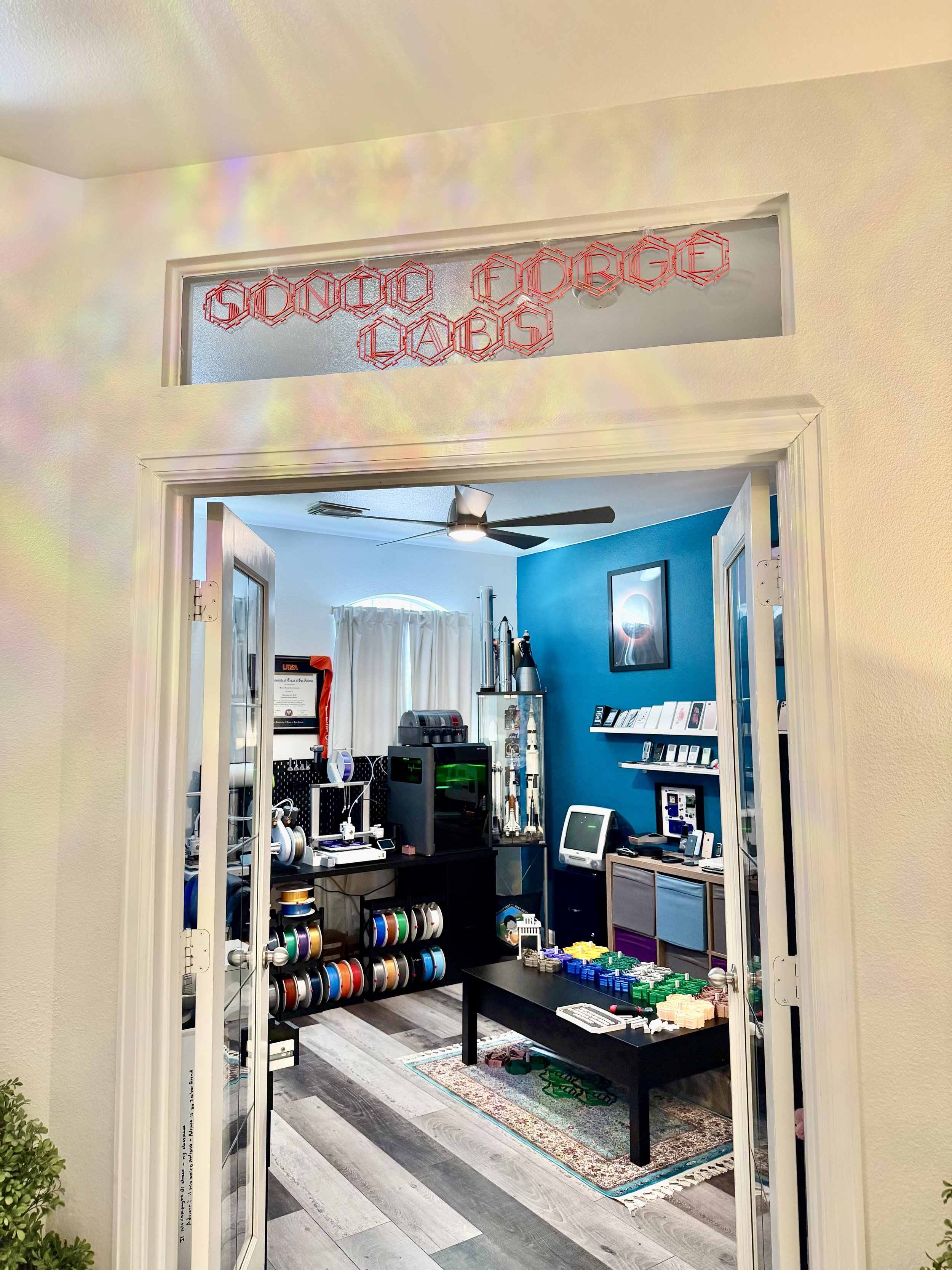 A room filled with various electronic and gaming equipment, including shelves with computer parts, controllers, and cables, with a bright blue wall and a ceiling fan.