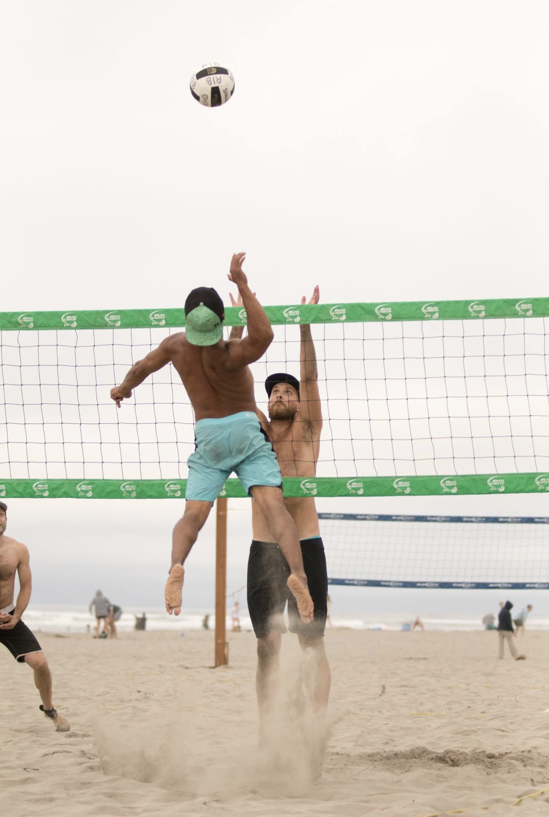 Beach volleyball players jumping at the net, one in mid-air hitting the ball, beach and cloudy sky in background.