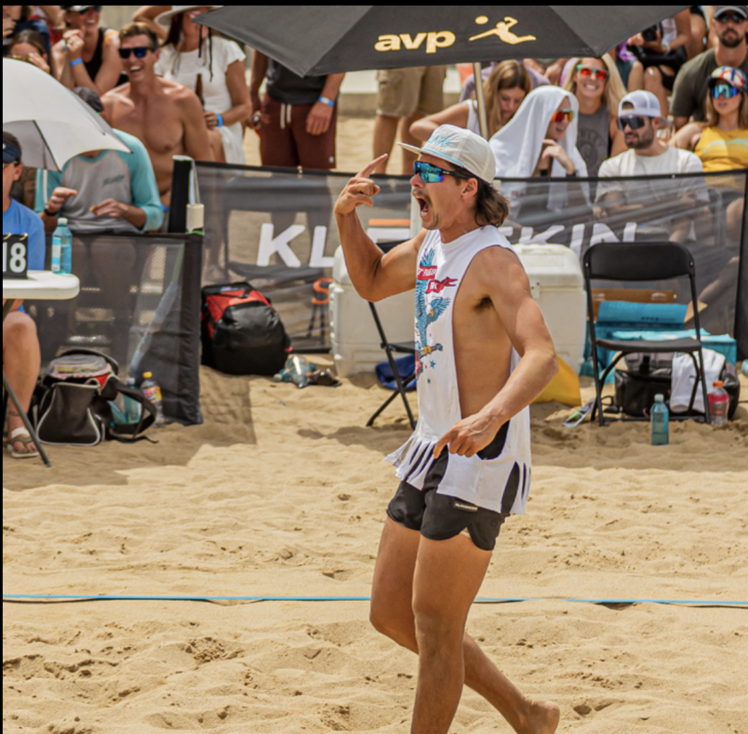 A man in beachwear, sunglasses, and a cap, gesturing animatedly during a beach volleyball game with a crowd watching behind a black barrier.