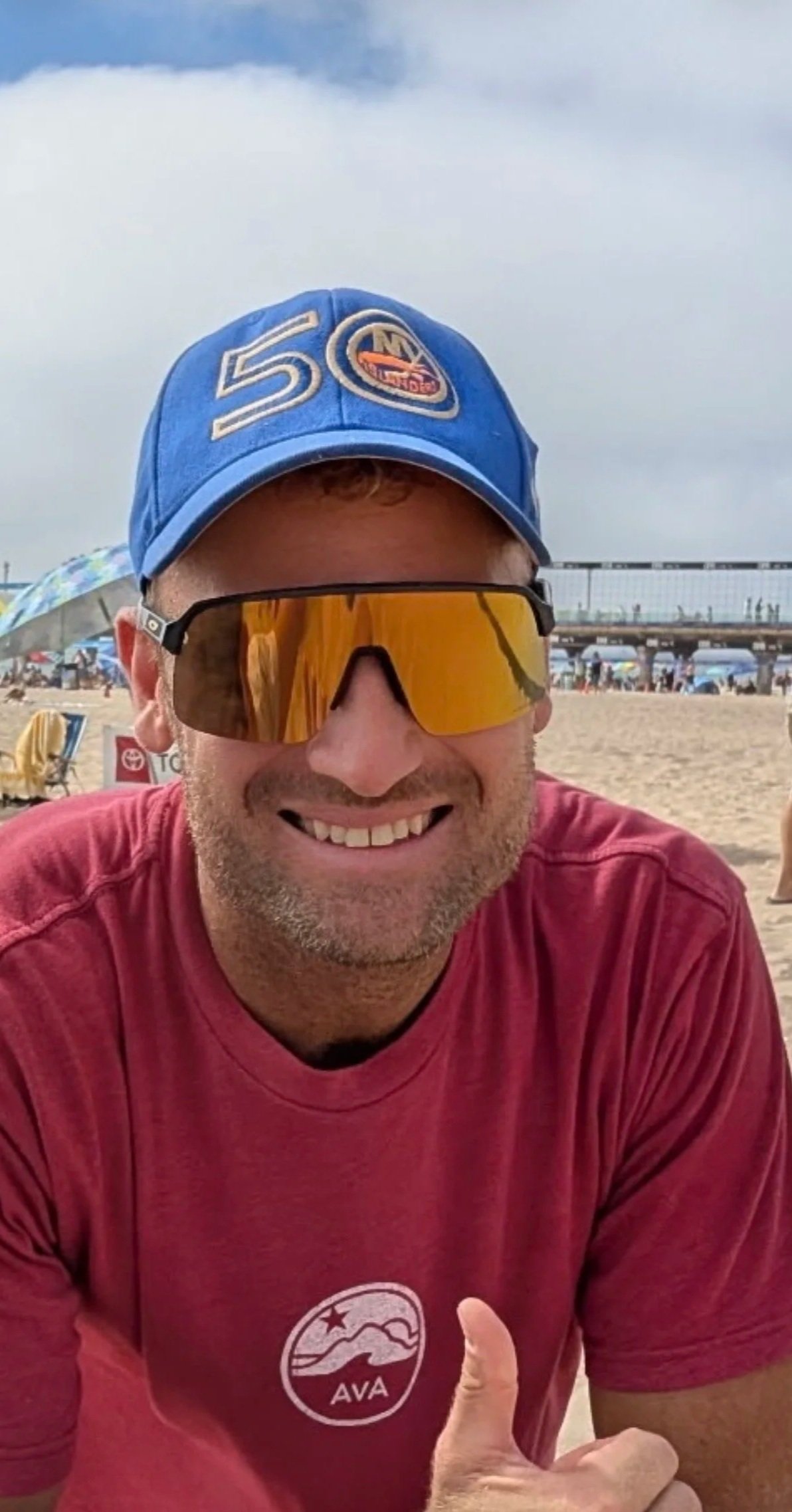 A man smiling and giving a thumbs-up, wearing sunglasses and a blue cap with a 50th logo, sitting on a sandy beach with umbrellas and people in the background.