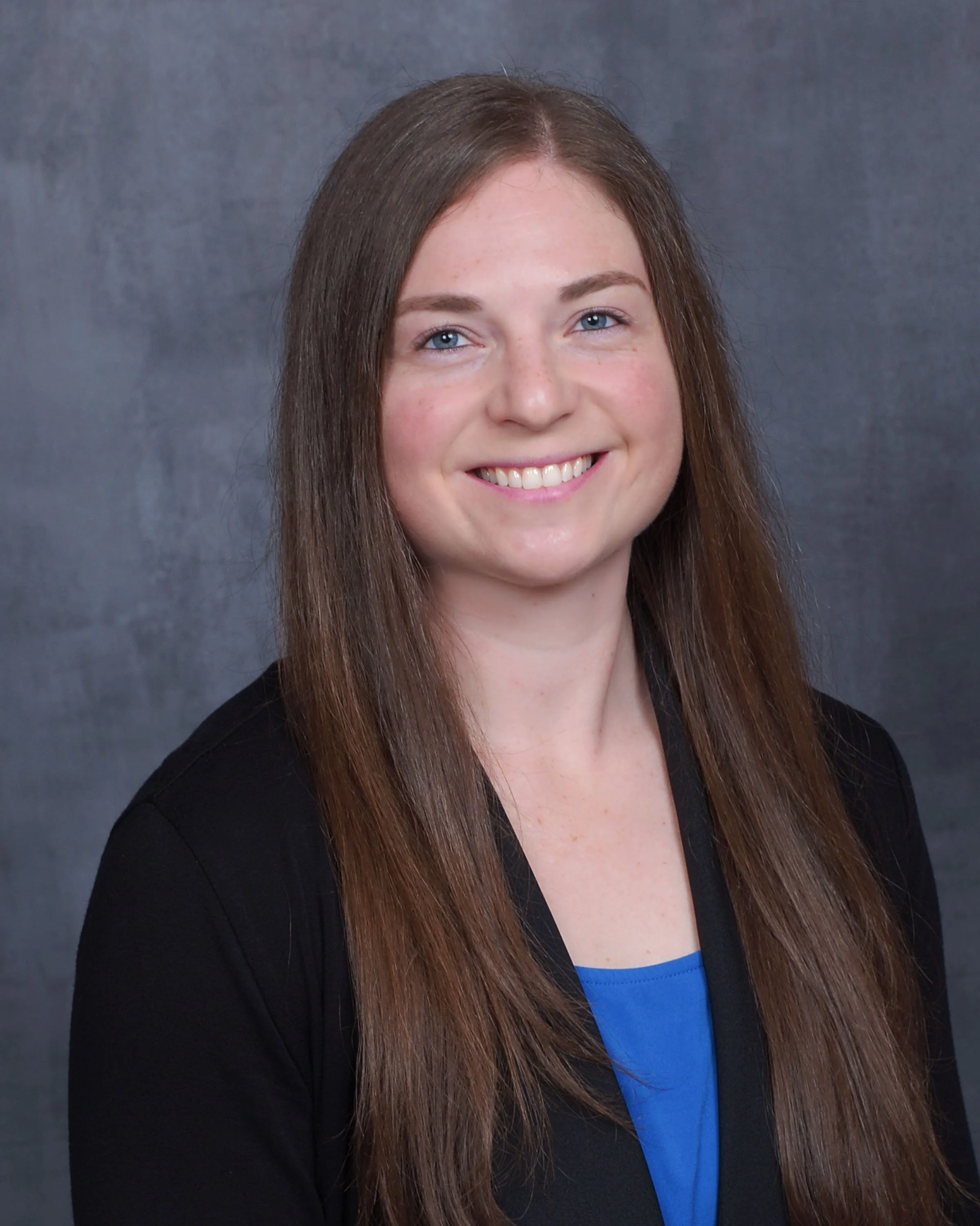 Headshot of a young woman with long brown hair, fair complexion, and blue eyes, smiling, wearing a black blazer and a blue top, against a gray background.