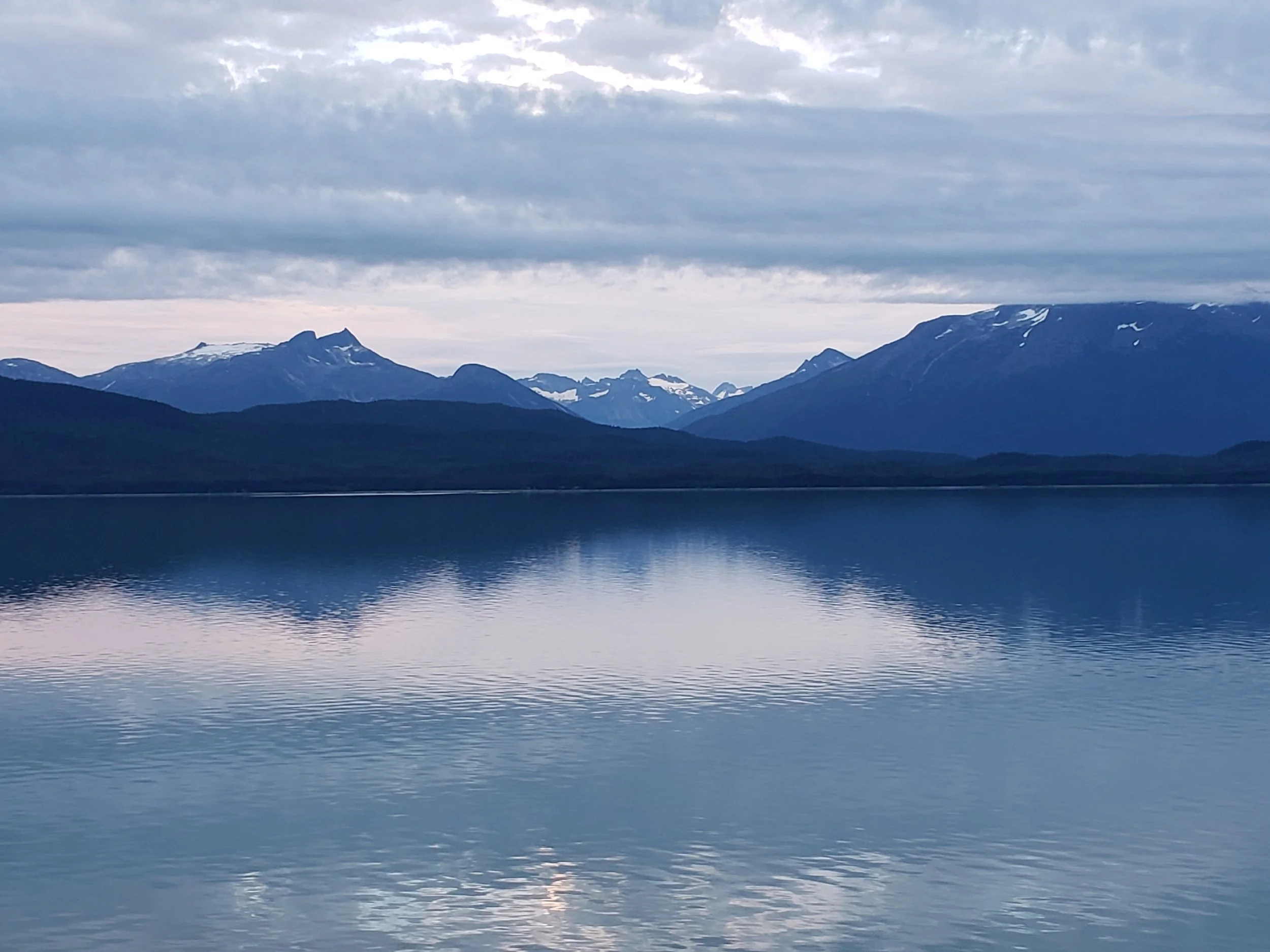 Scenic view of snow-capped mountains behind a calm body of water under a cloudy sky in Alaska.