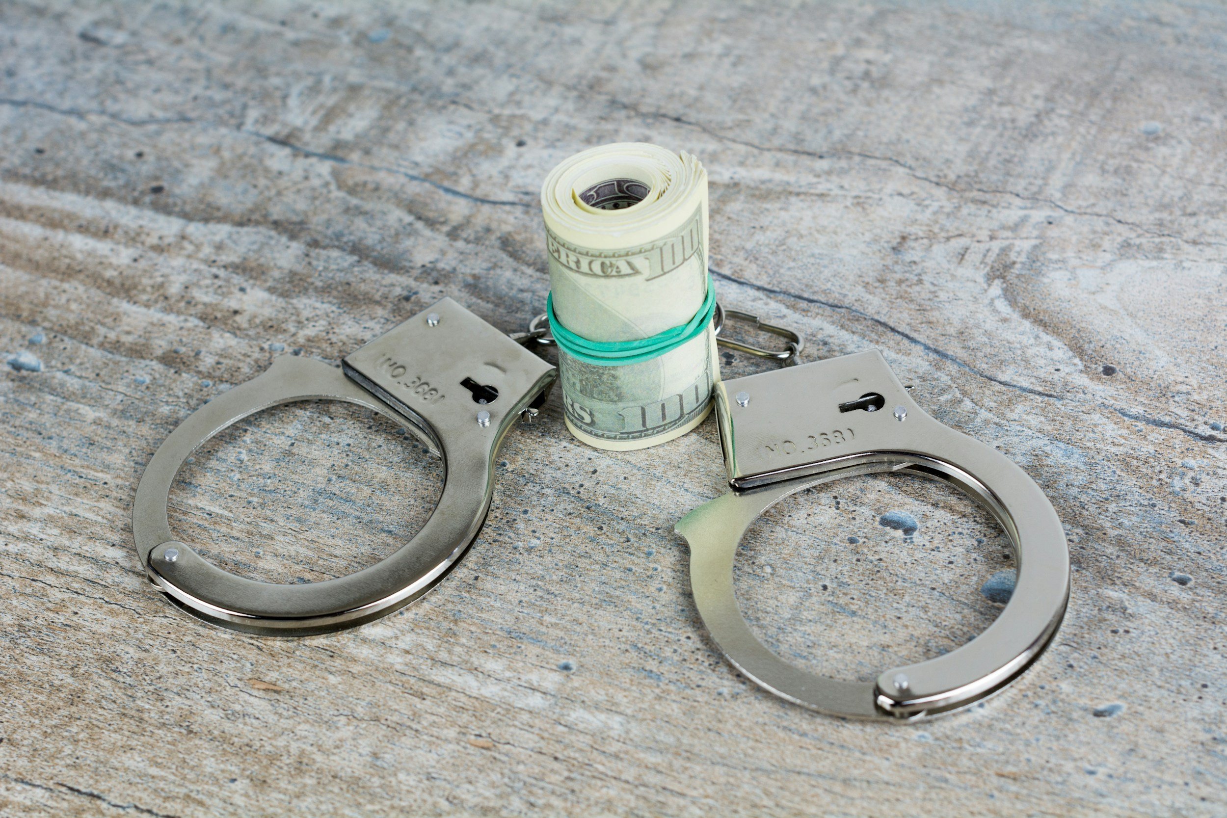 A rolled-up stack of US dollar bills secured with a green rubber band, placed inside a pair of metal handcuffs on a concrete surface.