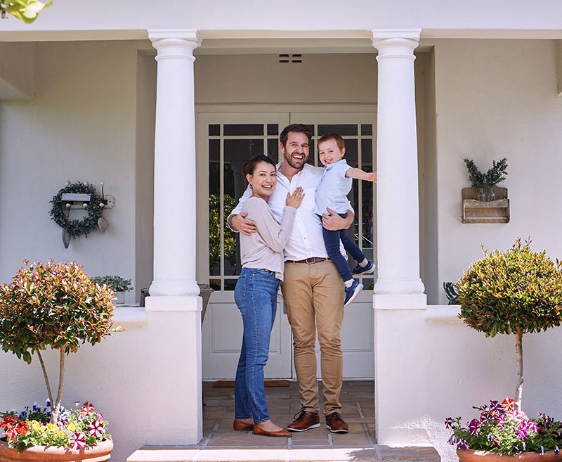 A happy family of three standing on the porch of a house. The man is holding a young boy, and the woman is standing next to them, smiling at the camera. The porch has two columns and potted plants and flowers.
