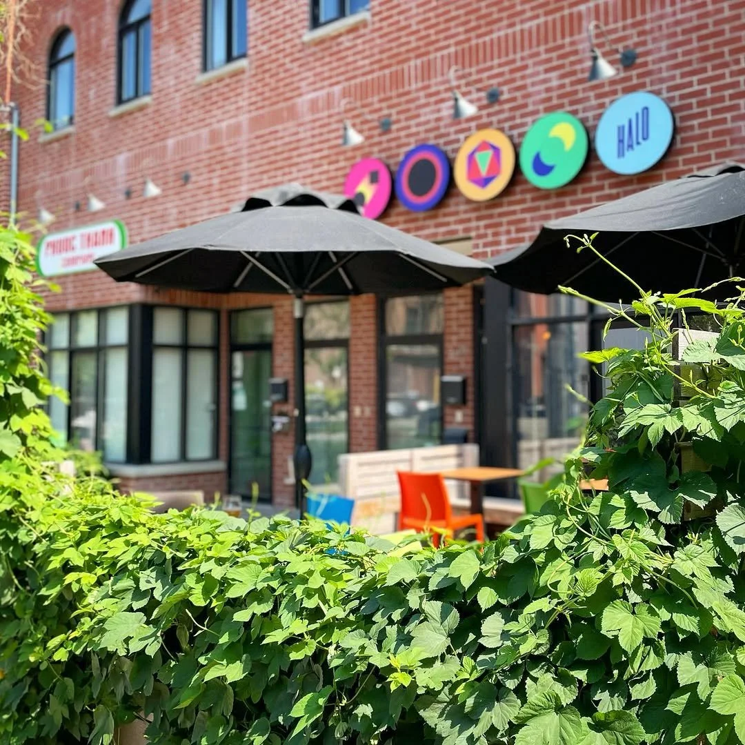 Outdoor patio with black umbrellas, colorful chairs, and a brick building with a sign and colorful circular decorations.
