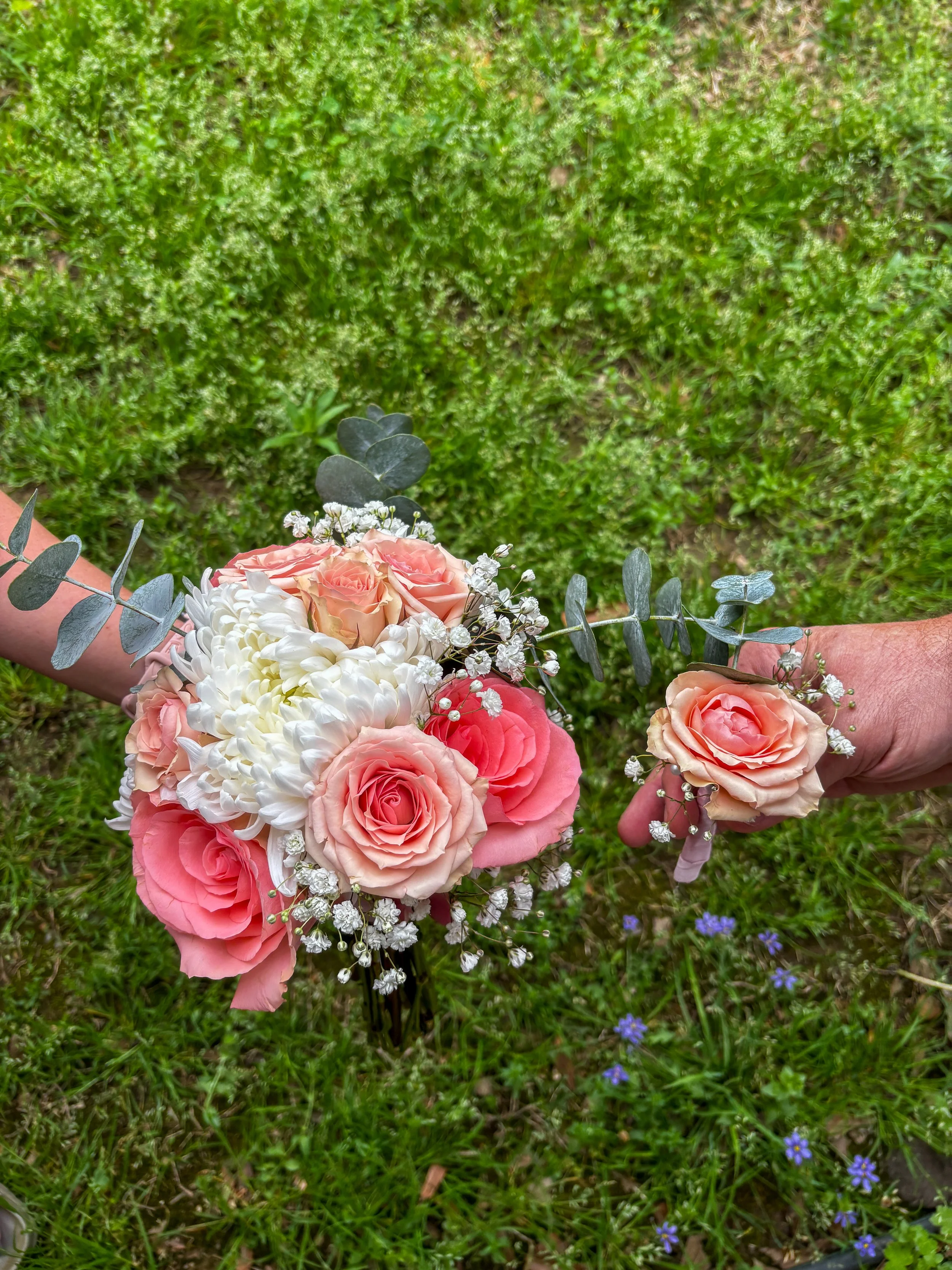 A person holding a bouquet of pink roses, small white flowers, and eucalyptus leaves, with a green grassy background.