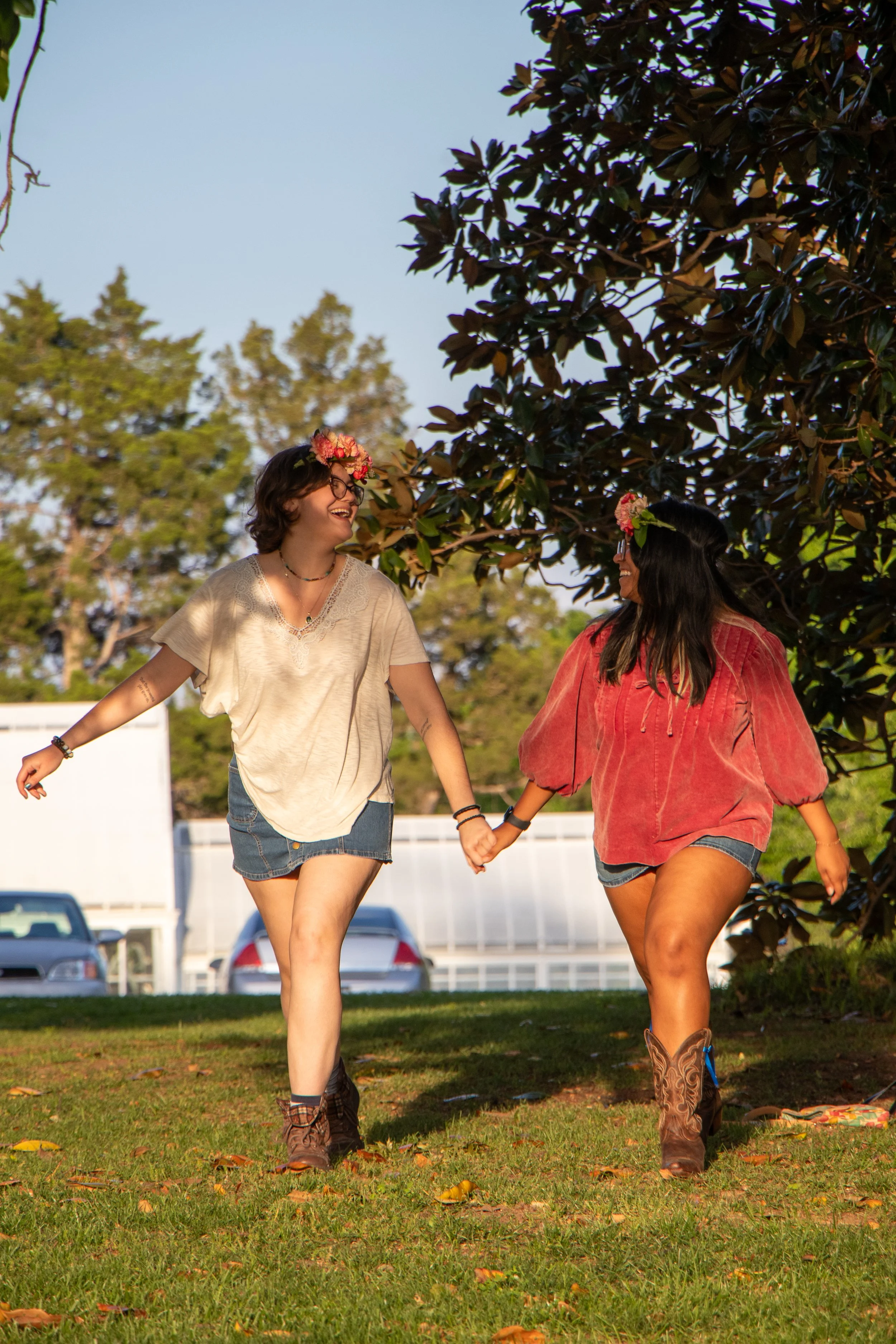 Two women holding hands and smiling while walking outdoors on a sunny day. One woman has short curly hair, glasses, a flower crown, and a cream-colored top with a denim skirt. The other woman has long dark hair, a flower crown, an orange-red top, and denim shorts. They are on a grassy area with trees and parked cars in the background.