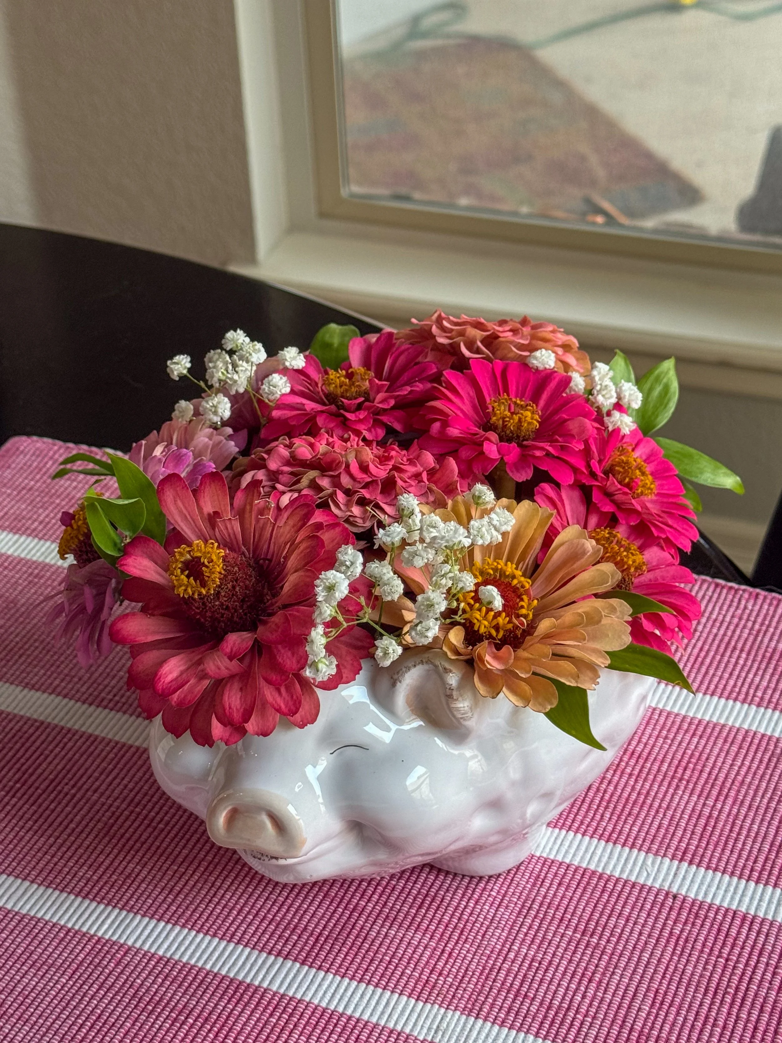 A bouquet of pink and orange flowers with green leaves in a white pig-shaped ceramic vase on a pink striped tablecloth.