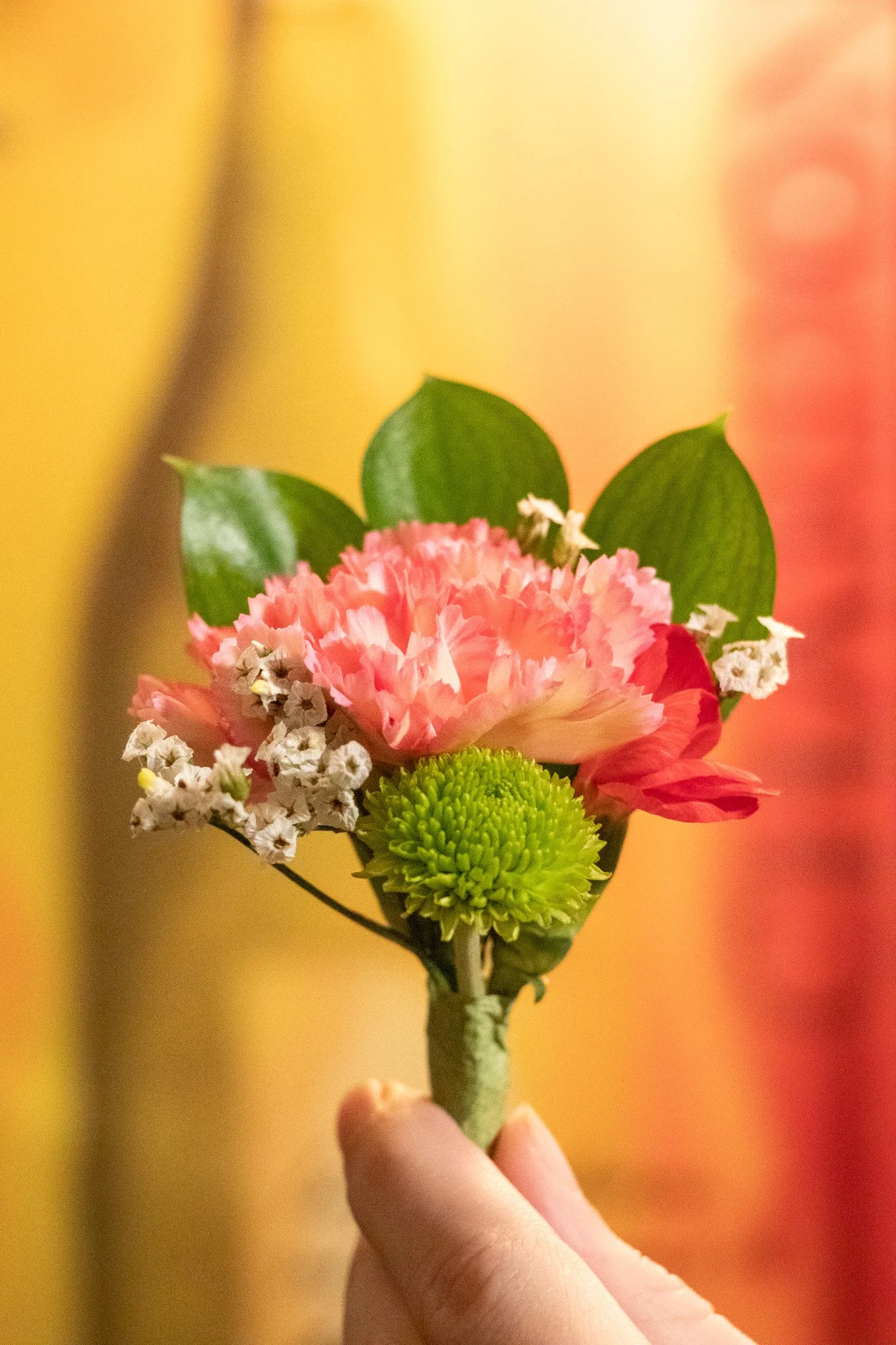 A person holding a small bouquet of mixed flowers with pink, white, green, and yellow flowers against a blurred yellow and orange background.