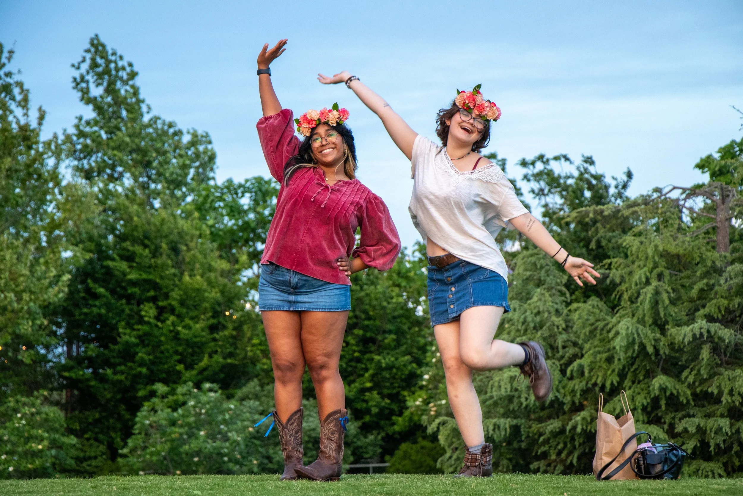 Two women with flower crowns jumping outdoors on a grassy field with trees in the background.