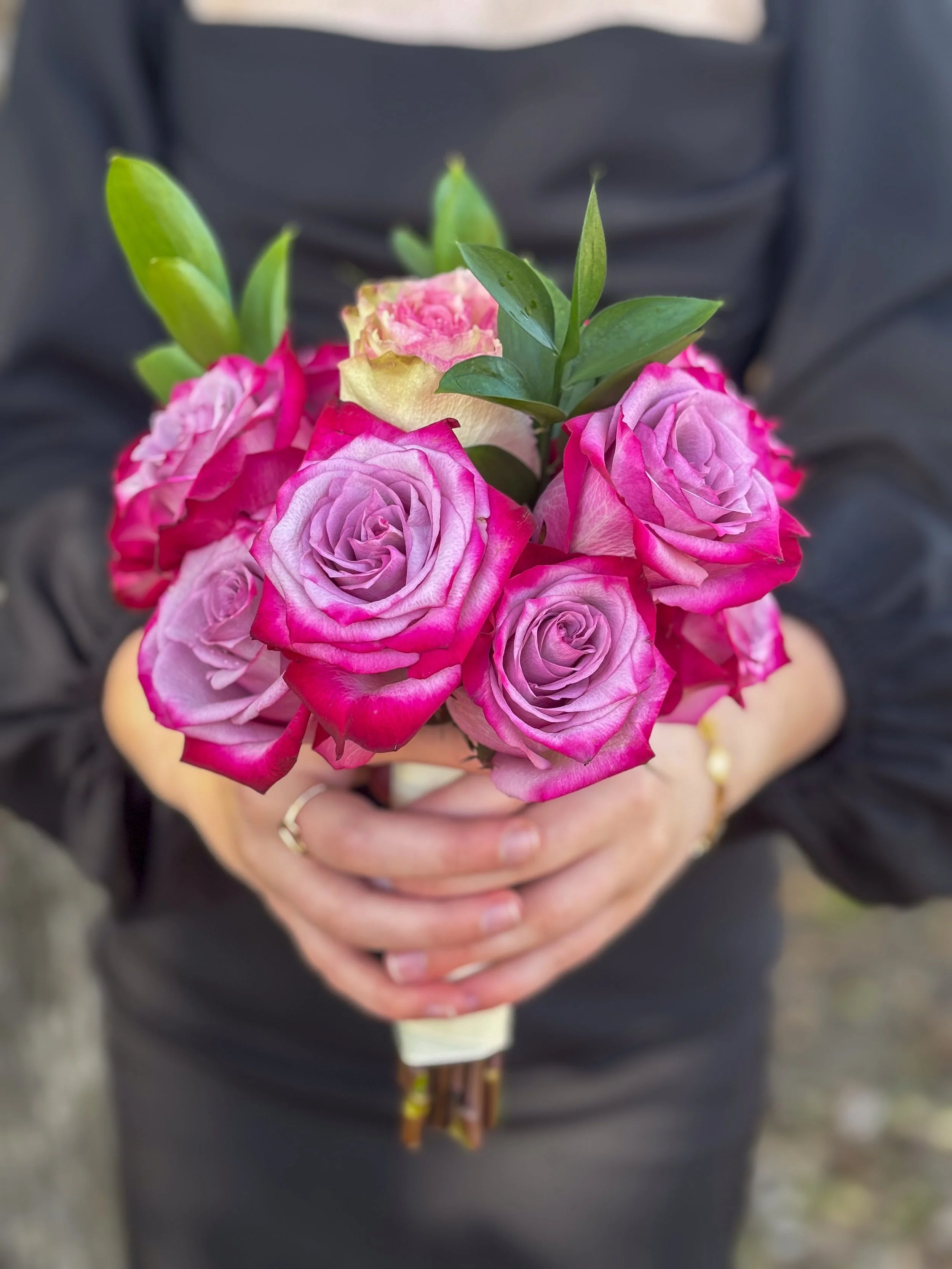 Person holding a bouquet of pink and purple roses with green leaves.