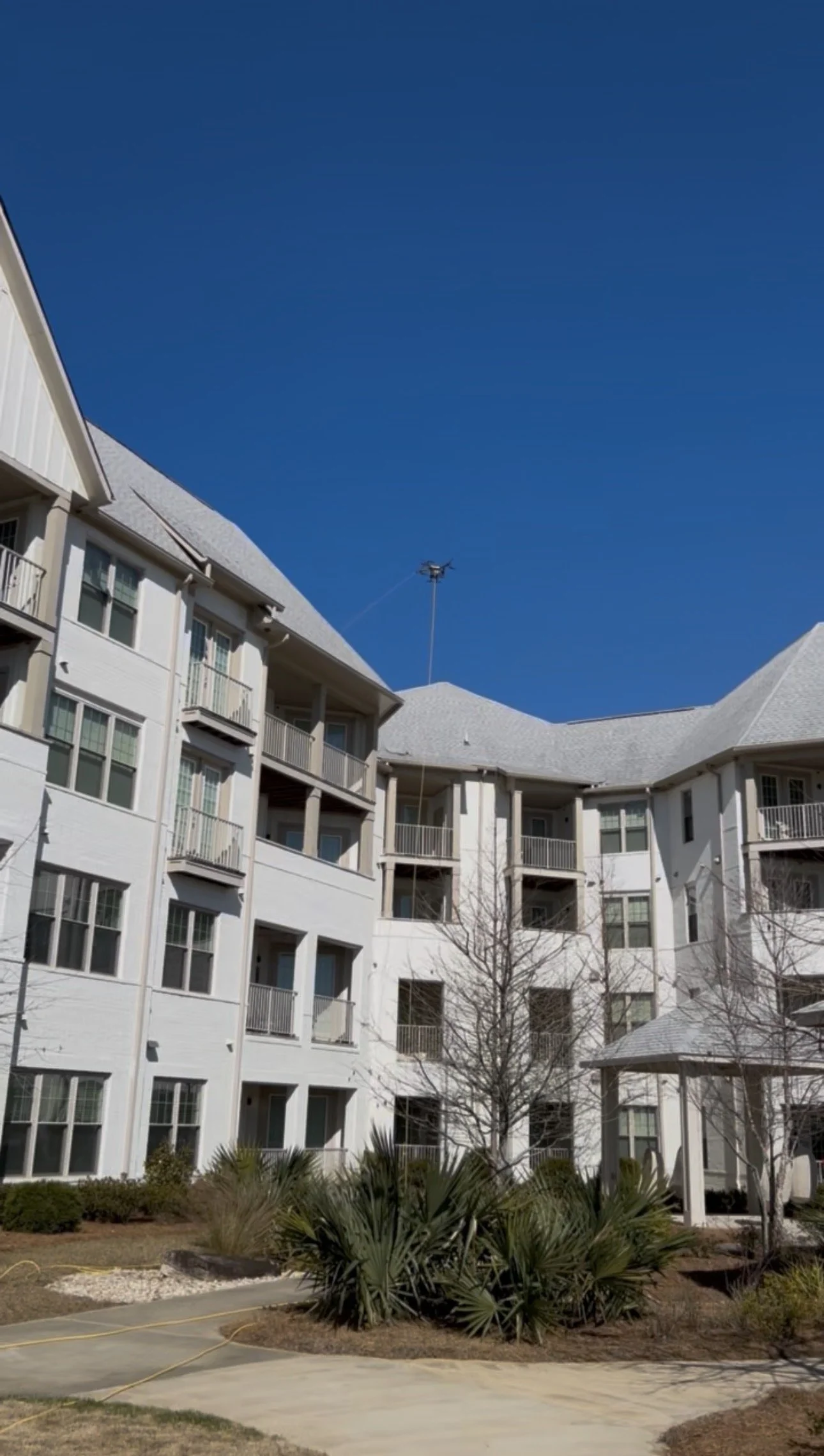 White multi-story apartment building with balconies against a bright blue sky, surrounded by desert plants and trees.