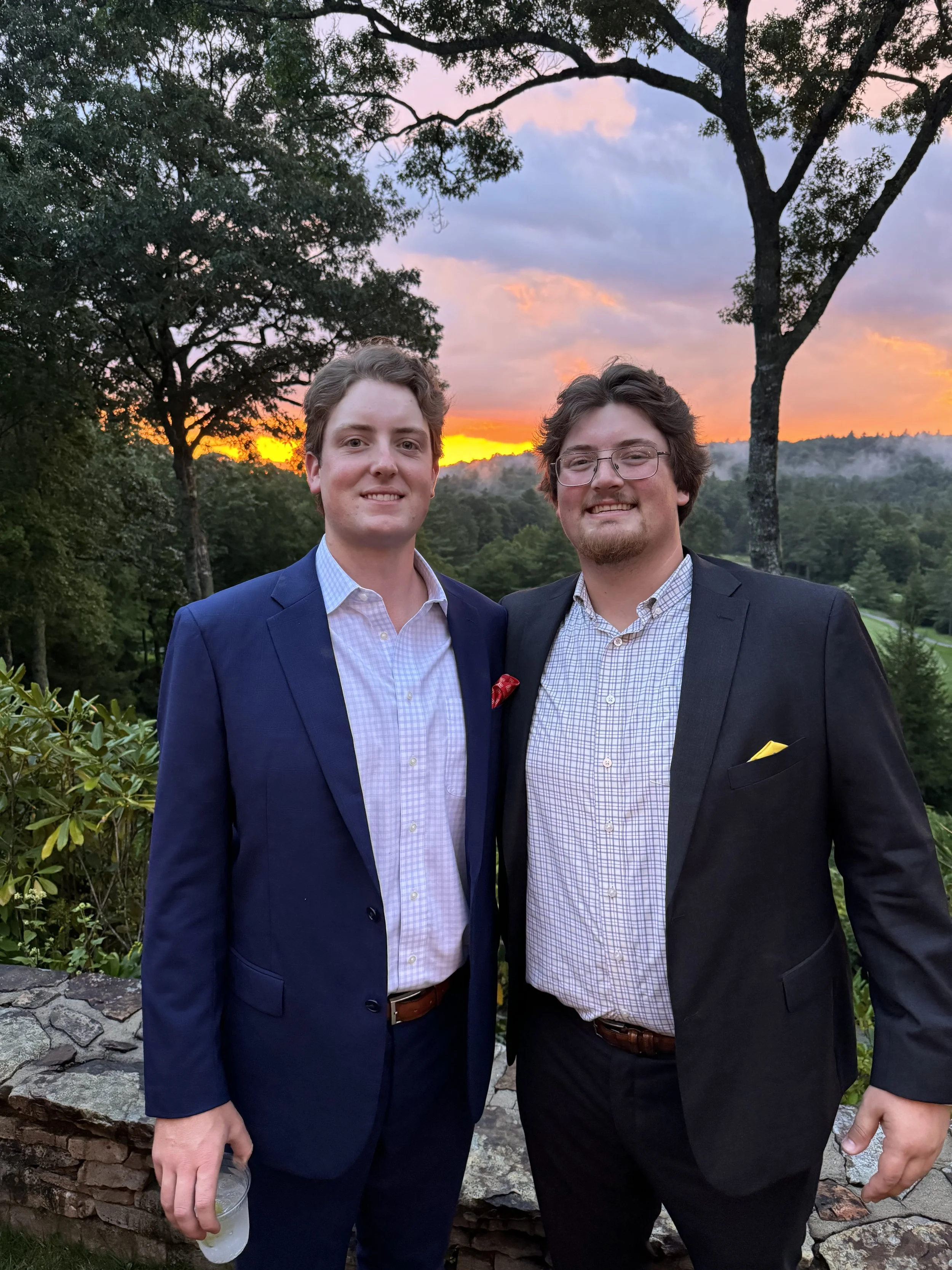 Two young men in suits standing outdoors during sunset with trees and a colorful sky in the background.